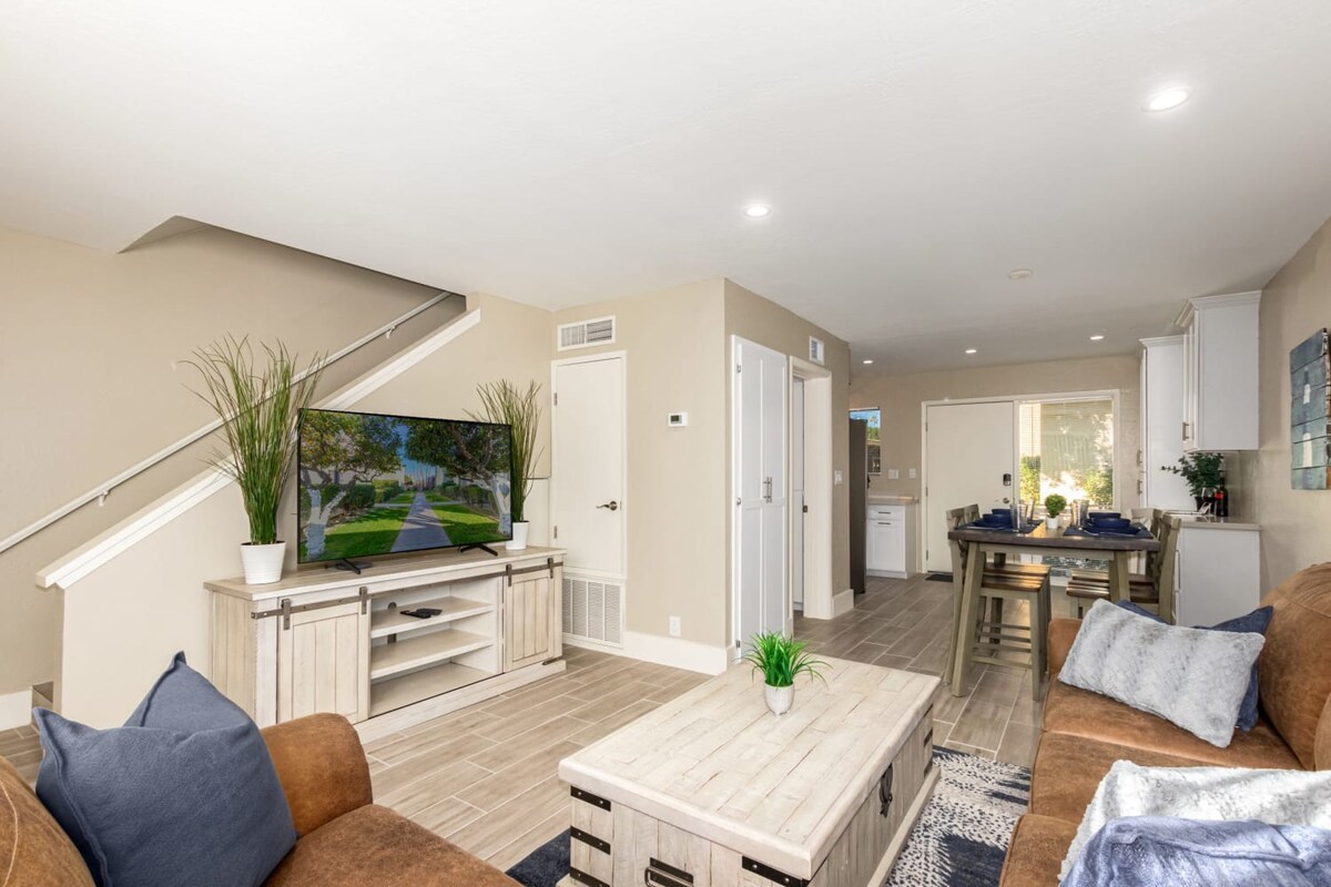 A spacious living area features a supple leather sofa and a loveseat, accompanied by a coffee table that doubles as a workspace. A 60" Smart TV is positioned for viewing, and natural light floods the room from the adjacent windows, creating a warm and inviting environment.