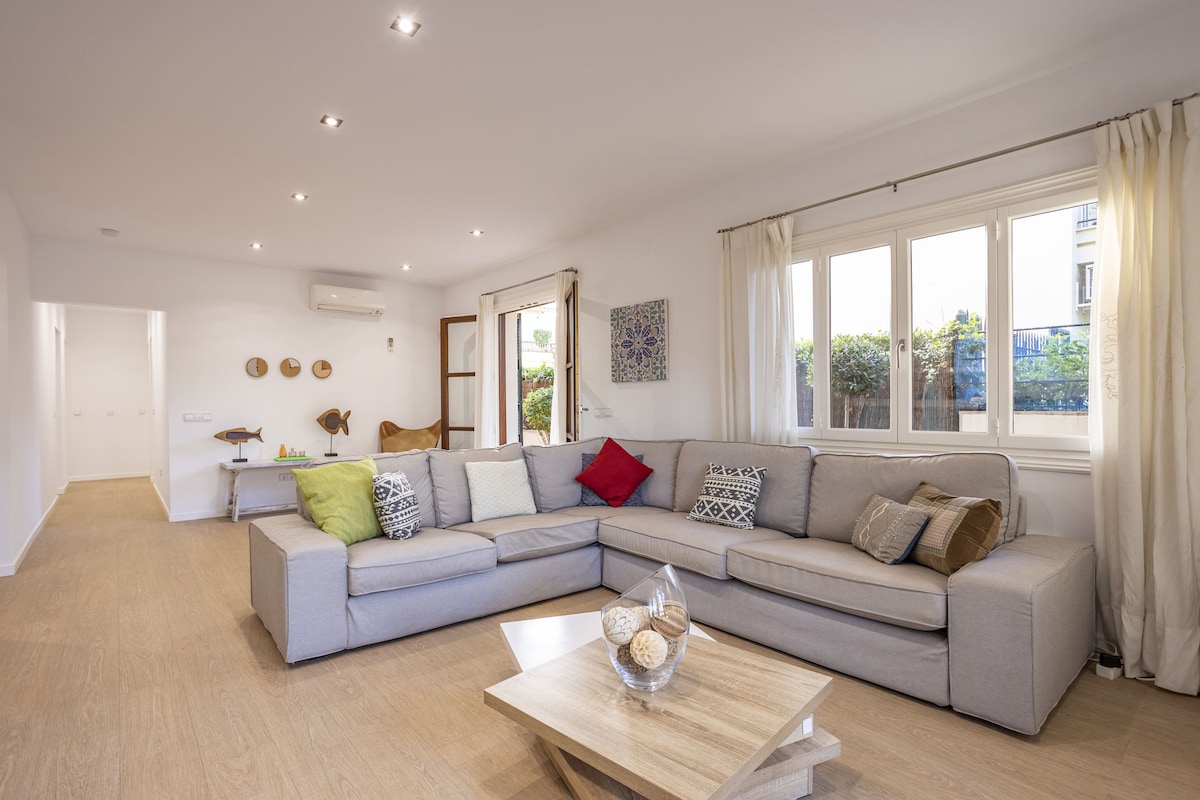 A spacious living area features light wood flooring and large windows that allow natural light to fill the room. A comfortable sectional sofa is arranged around a low wooden coffee table. Decorative cushions add color, while wall art accents the neutral walls. Air conditioning is visible.