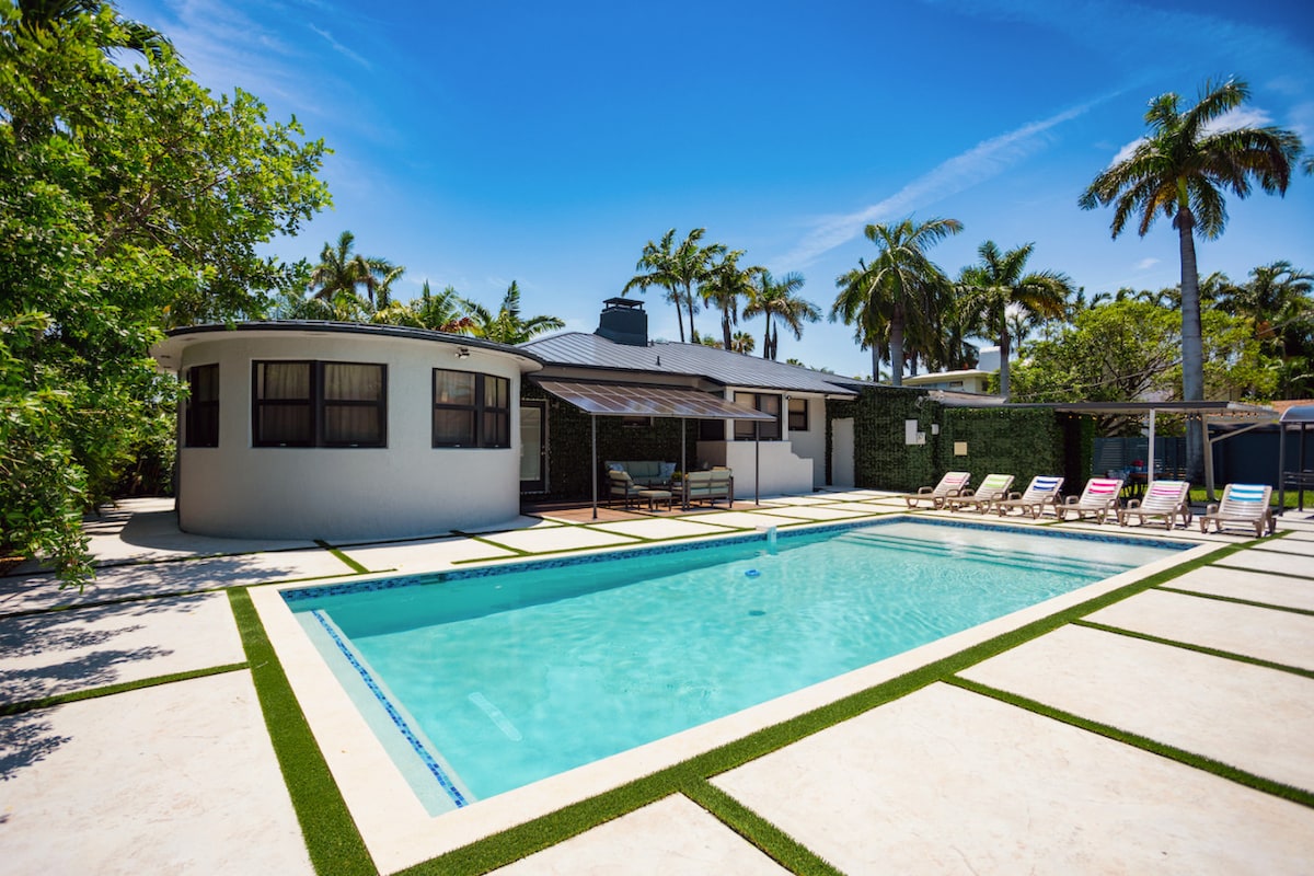 An inviting outdoor space features a newly constructed heated pool surrounded by landscaped gardens. A lounge area with sun chairs and shade canopies is positioned beside the pool, complementing the villa's modern architecture and palm trees in the background.