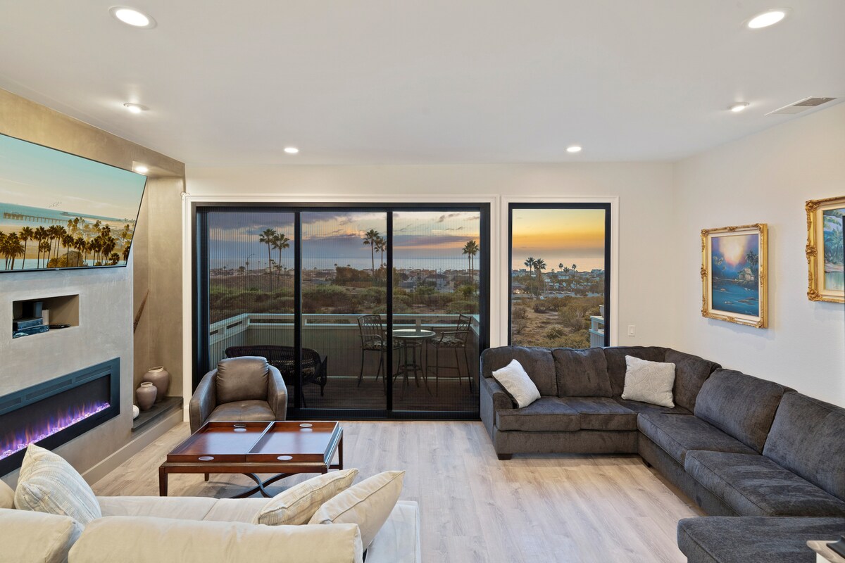 A spacious living room features large glass doors that offer stunning ocean views at sunset. A comfortable sectional sofa is positioned around a coffee table, with a fireplace visible on the side. Subtle artwork and decor add to the inviting ambiance.