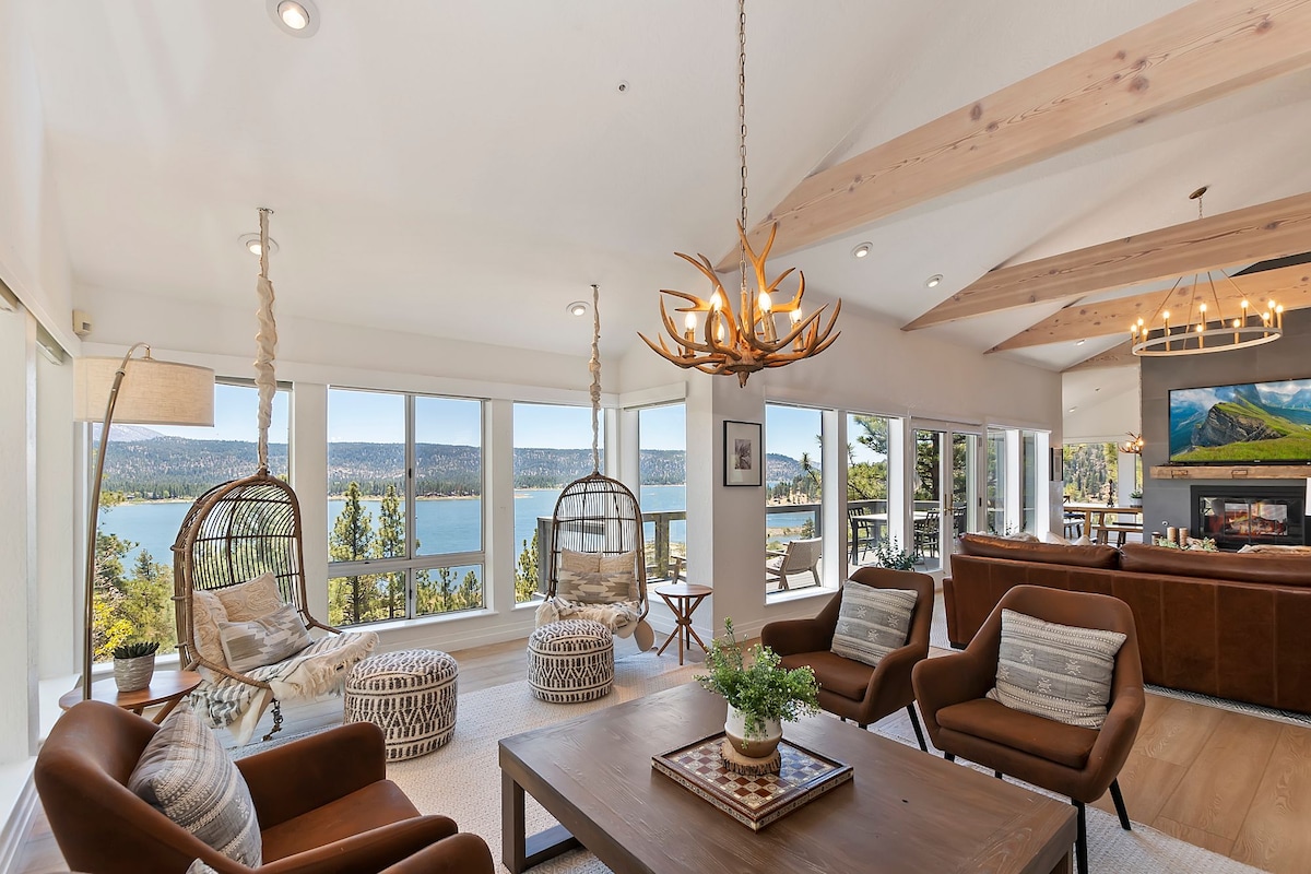 A bright living area showcases large windows framing expansive views of the lake. Two hanging lounge chairs with woven details provide a cozy seating option. A wooden table is centered between two modern armchairs, while a chandelier made of antlers adds a rustic touch to the space.