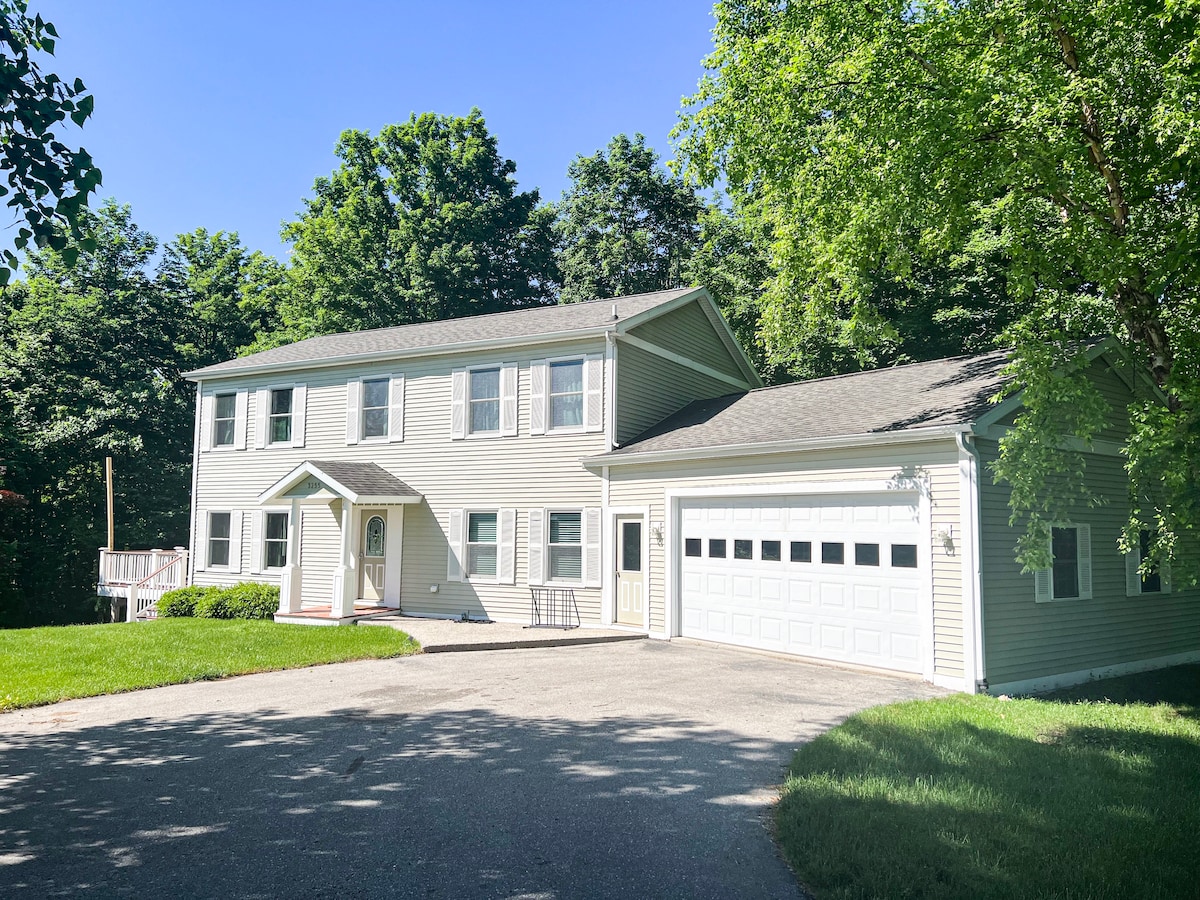 A two-story home is surrounded by lush green trees, featuring a light-colored exterior with multiple windows and an inviting front entrance. A spacious driveway leads to a garage, offering easy access. The well-maintained lawn is complemented by natural landscaping.