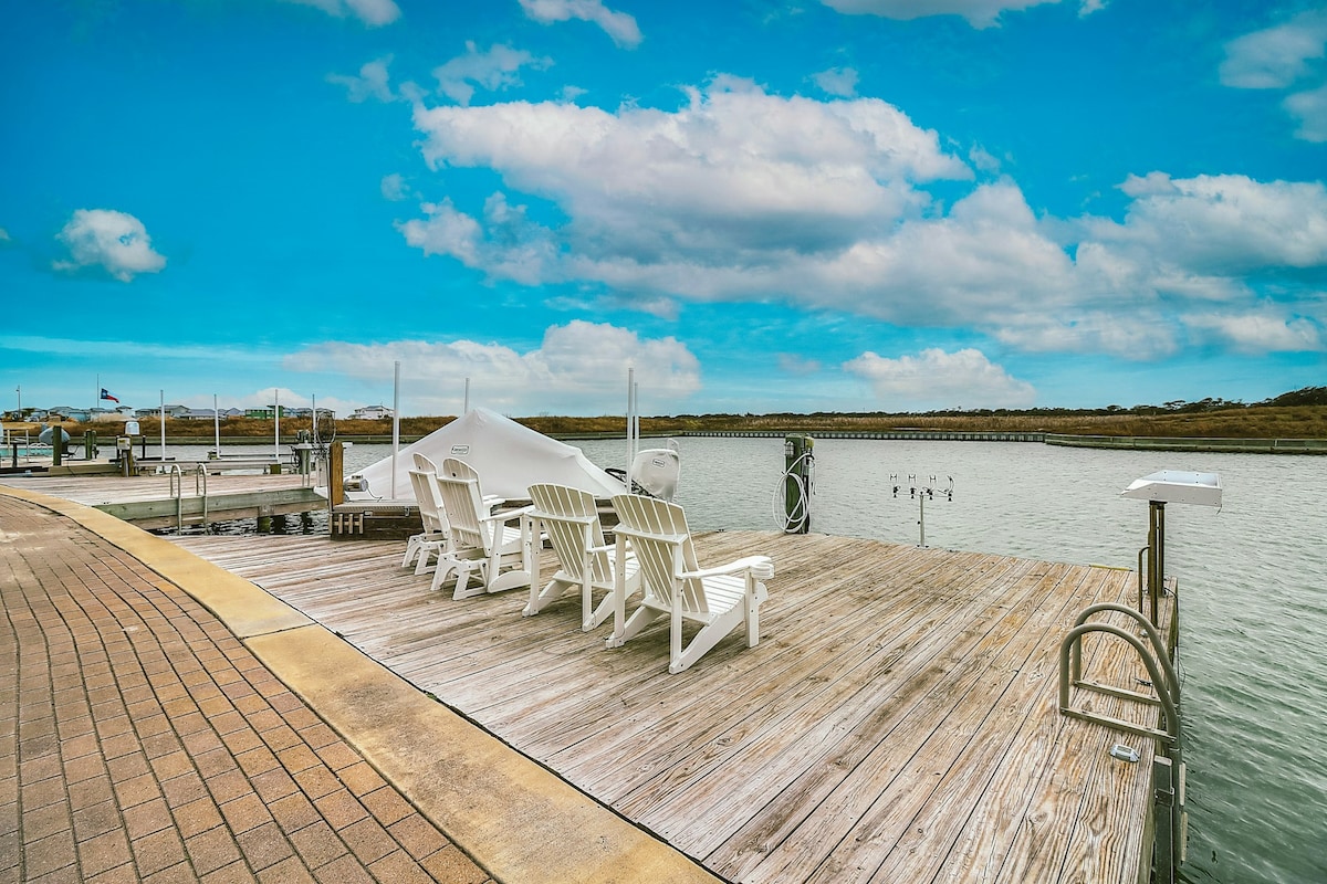 A private dock features several white Adirondack chairs arranged for relaxation. The wooden surface extends over the water, surrounded by greenery and an expansive sky, creating an inviting spot for enjoying the serene waterfront views.
