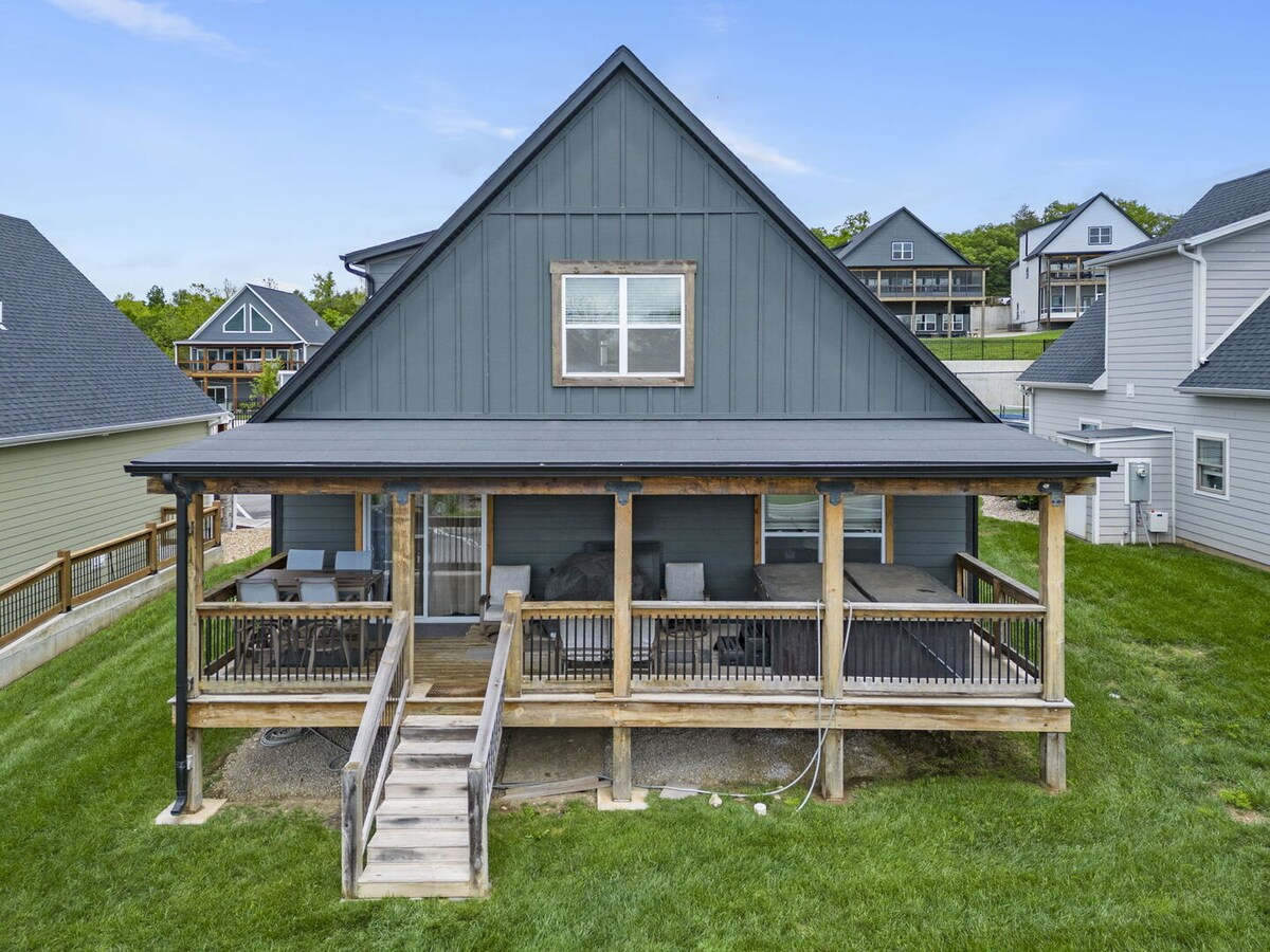 The exterior of Cove Cottage features a welcoming grey facade with a peaked roof. A spacious porch is observed, surrounded by a wooden railing, providing versatility for outdoor relaxation. Green grass frames the structure, enhancing the inviting ambiance of the outdoor space.