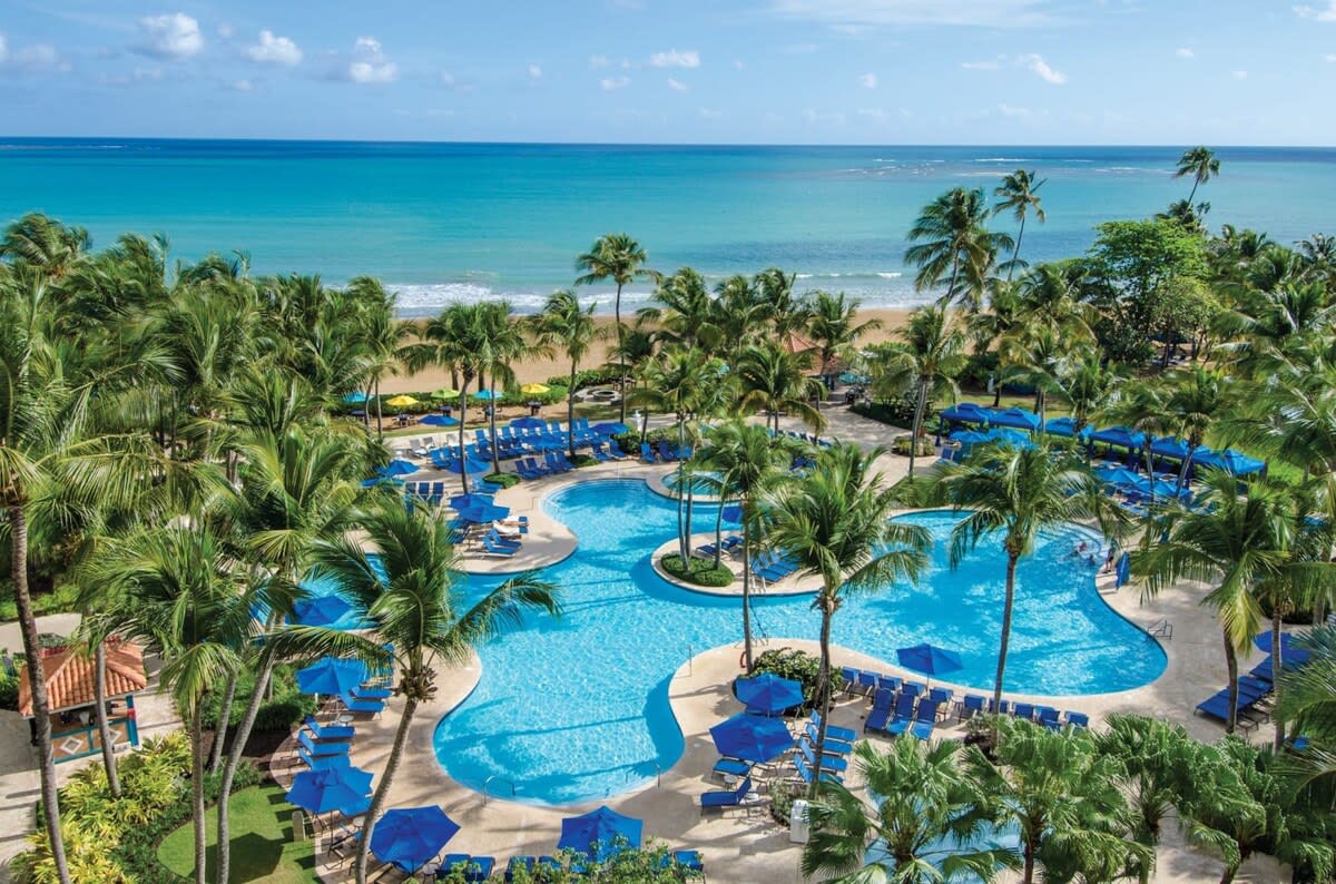 An inviting pool area features multiple interconnected pools surrounded by lush palm trees. Bright blue loungers and umbrellas are scattered around the pools, offering comfortable seating options. The expansive beach is visible in the background, blending into the turquoise water under a clear blue sky.