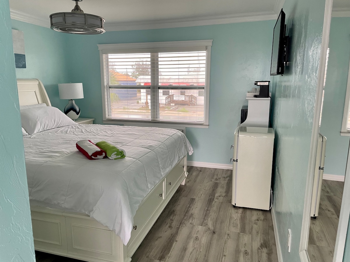 A well-lit studio room features a spacious king-size bed with a white quilt. A small kitchenette equipped with a mini fridge and microwave is visible. Natural light enters through two windows with blinds, and a contemporary pendant light hangs from the ceiling.