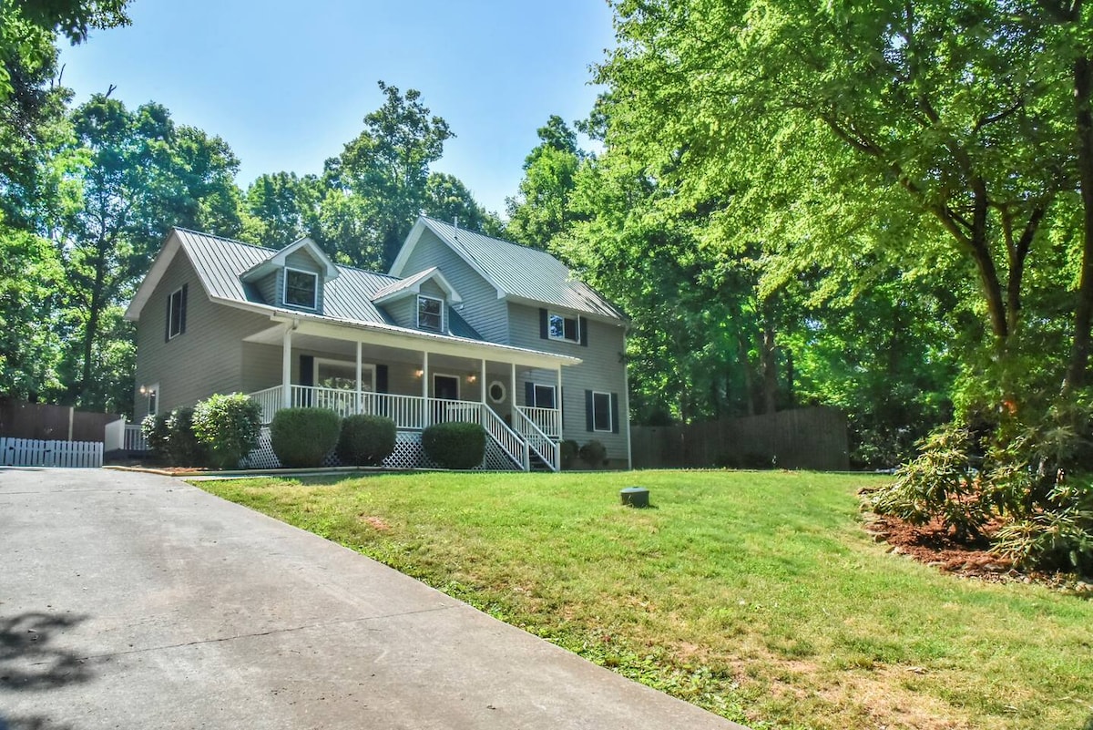 A spacious two-story retreat is framed by lush greenery, featuring a welcoming porch with steps leading up to the entrance. The well-maintained driveway is bordered by manicured lawns and shrubbery, creating a serene and inviting exterior.