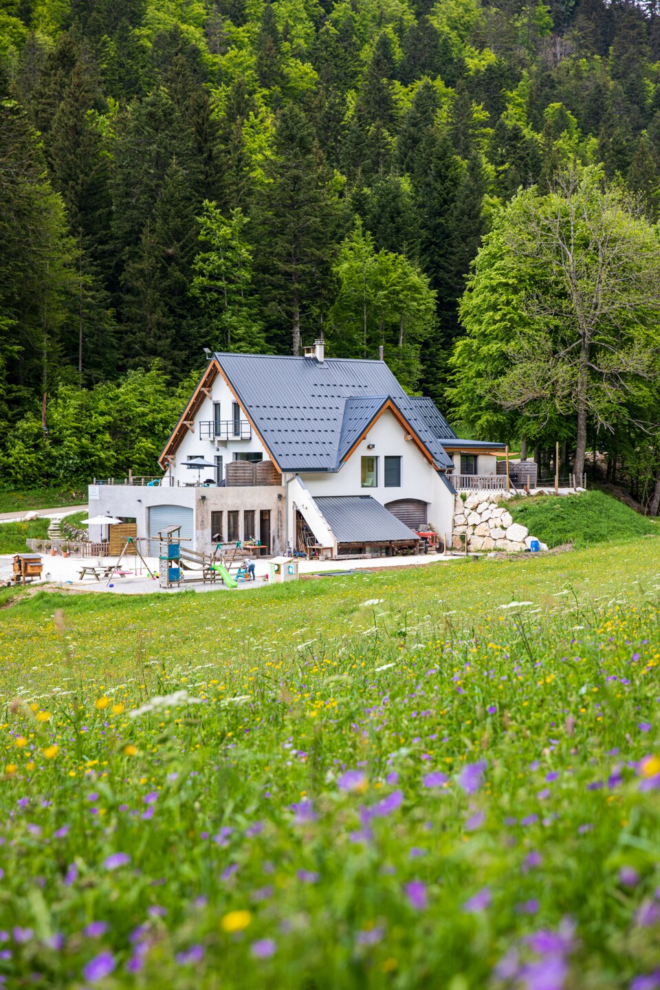 The guesthouse is set against a lush green backdrop, surrounded by trees. The architectural style combines modern elements with natural surroundings, featuring a sloped roof and a terrace area. An inviting outdoor space is visible, complete with seating and dining areas amidst colorful wildflowers.