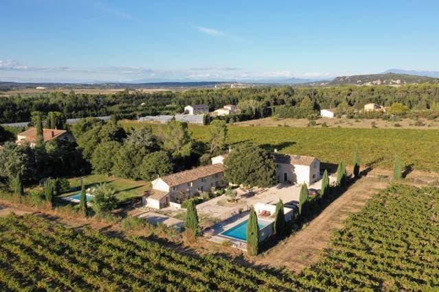 Aerial view of a property surrounded by vineyards, showcasing a central swimming pool and landscaped grounds. Nearby buildings feature terracotta roofs, with trees providing shade. Rolling hills and distant mountains are visible, indicating a serene rural setting.