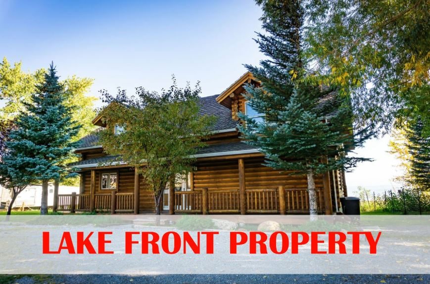 A charming log cabin is showcased, set against a backdrop of mature trees and landscaped grounds. The exterior features a combination of wood siding and a sloped roof. A welcoming porch invites guests to enjoy the tranquil lakefront setting.