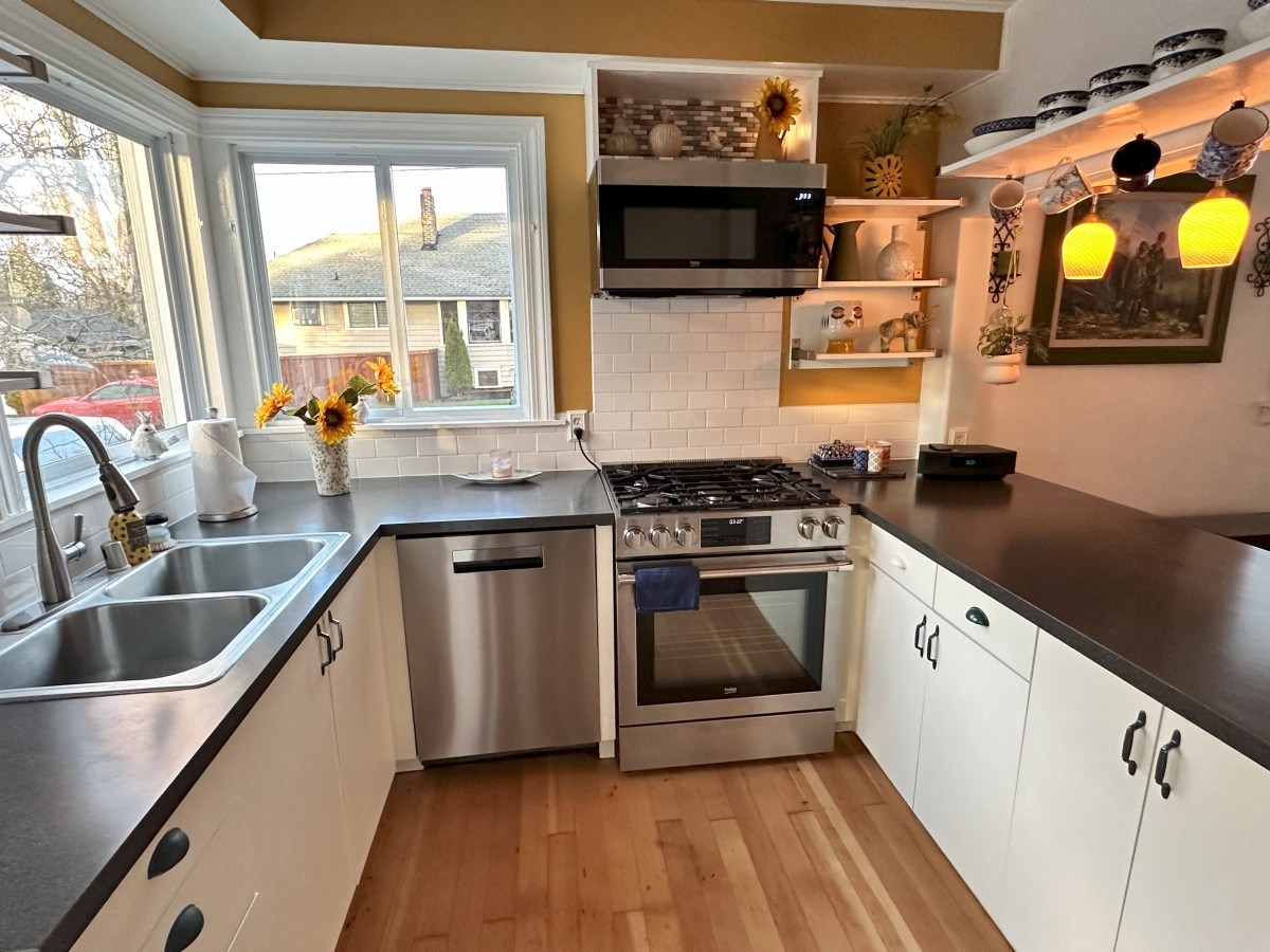 A bright kitchen is showcased with a large window allowing natural light to stream in. New stainless steel appliances, including a gas range and a dishwasher, are present. White cabinetry and a dark countertop create a modern contrast, complemented by decorative items on shelves.