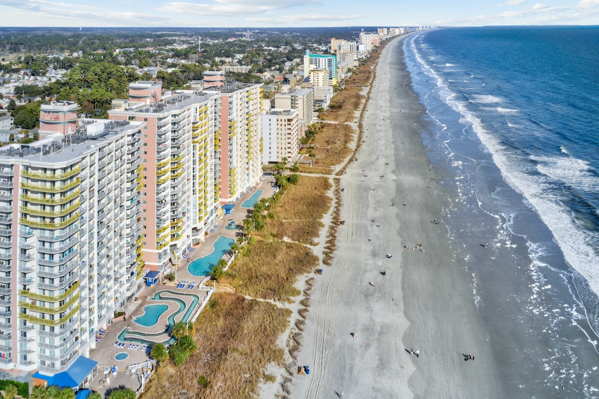 An aerial view showcases the oceanfront condo building along a sandy beach. The structure features multiple levels, with several pools visible below. The coastline extends into the distance, bordered by gentle waves meeting the shore, highlighting the expansive stretch of beach.