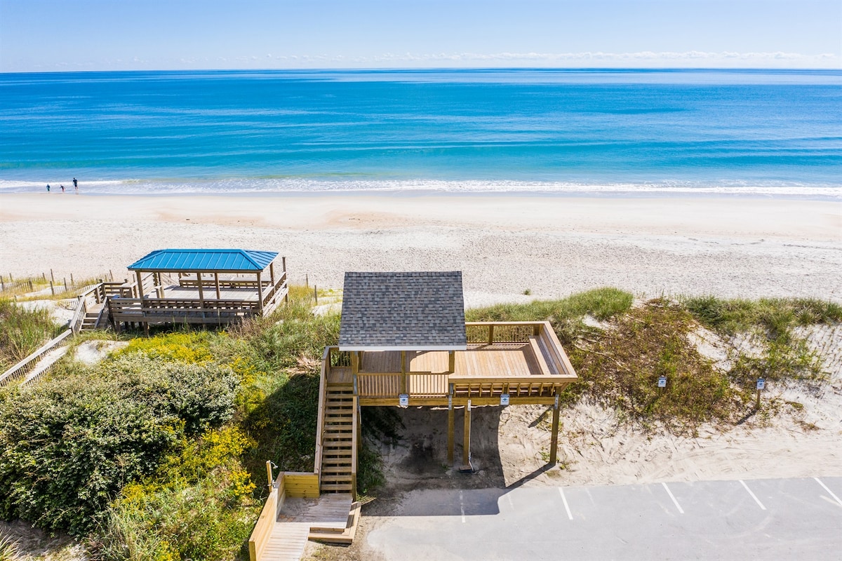A sandy coastline stretches toward the horizon, with gentle waves lapping at the shore. A wooden gazebo with a sloped roof stands close to the beach, providing a shaded area. A pathway leads from the gazebo to the beach, inviting relaxation by the ocean.