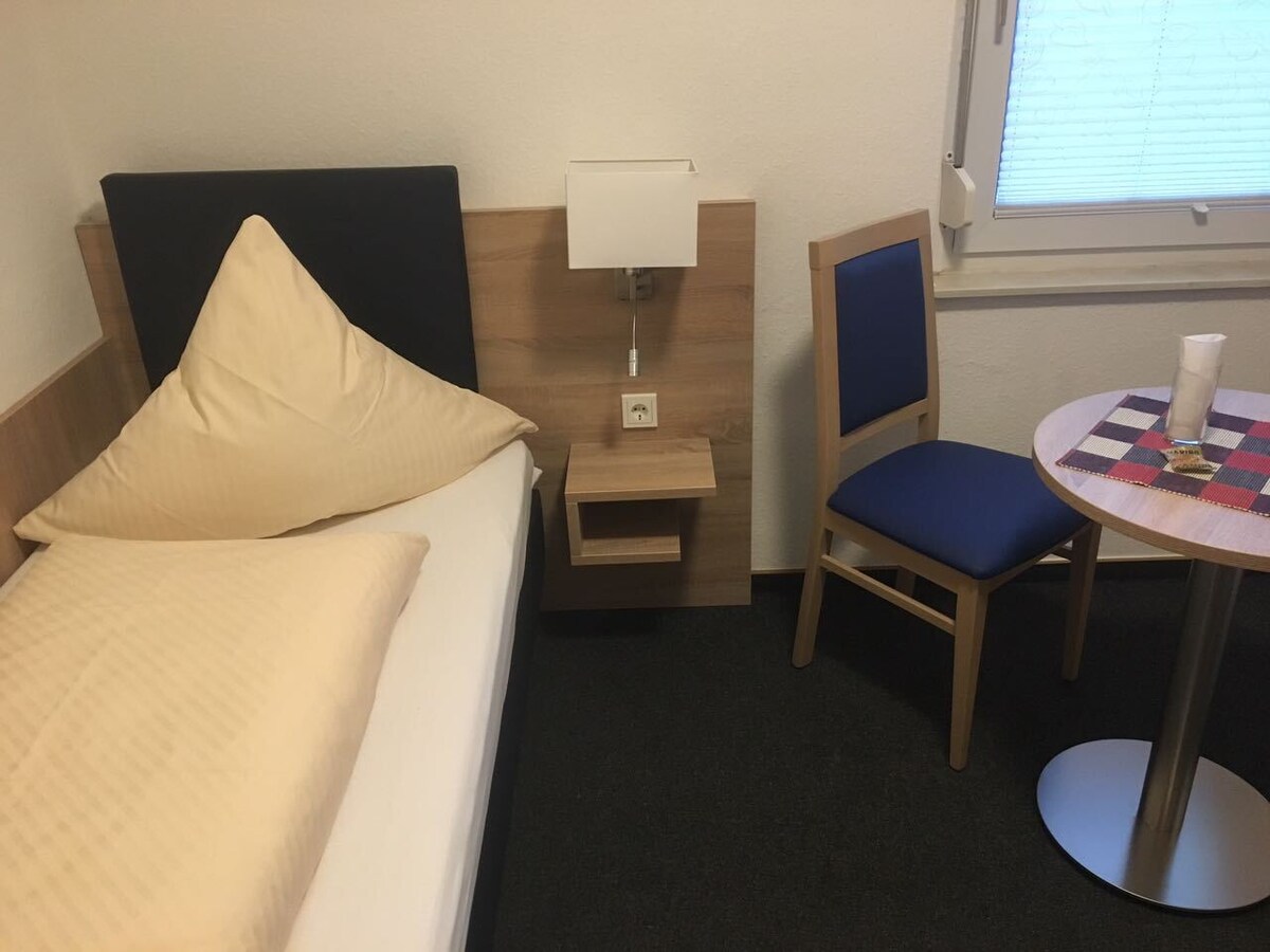 A compact room features a single bed with a petite decorative pillow and a small nightstand equipped with a lamp. A chair with blue upholstery is positioned beside a round table topped with a checkered mat. Natural light filters through the window with blinds.