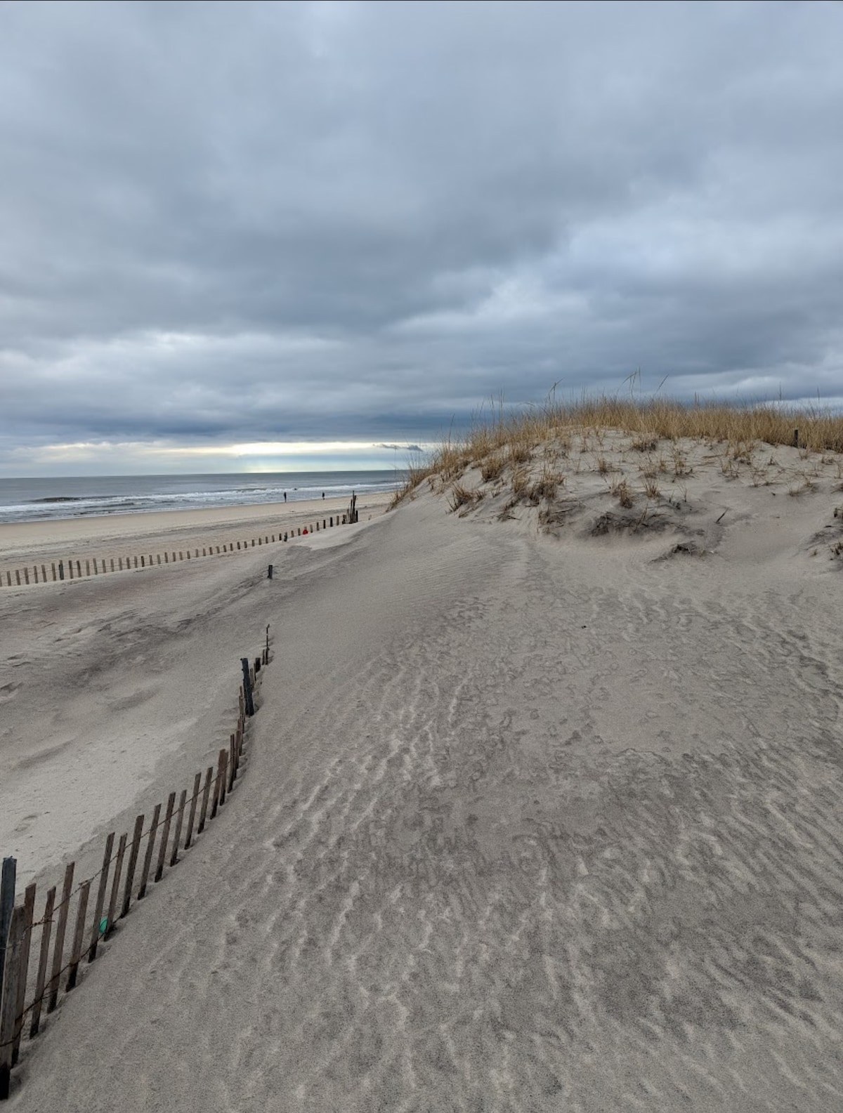 A sandy dune is depicted with patches of tall grass at its peak. A smooth surface leads down towards a beach, with gentle waves visible in the distance. Grey clouds give the scene a soft, muted light, while wooden fencing runs along the edges of the sand.