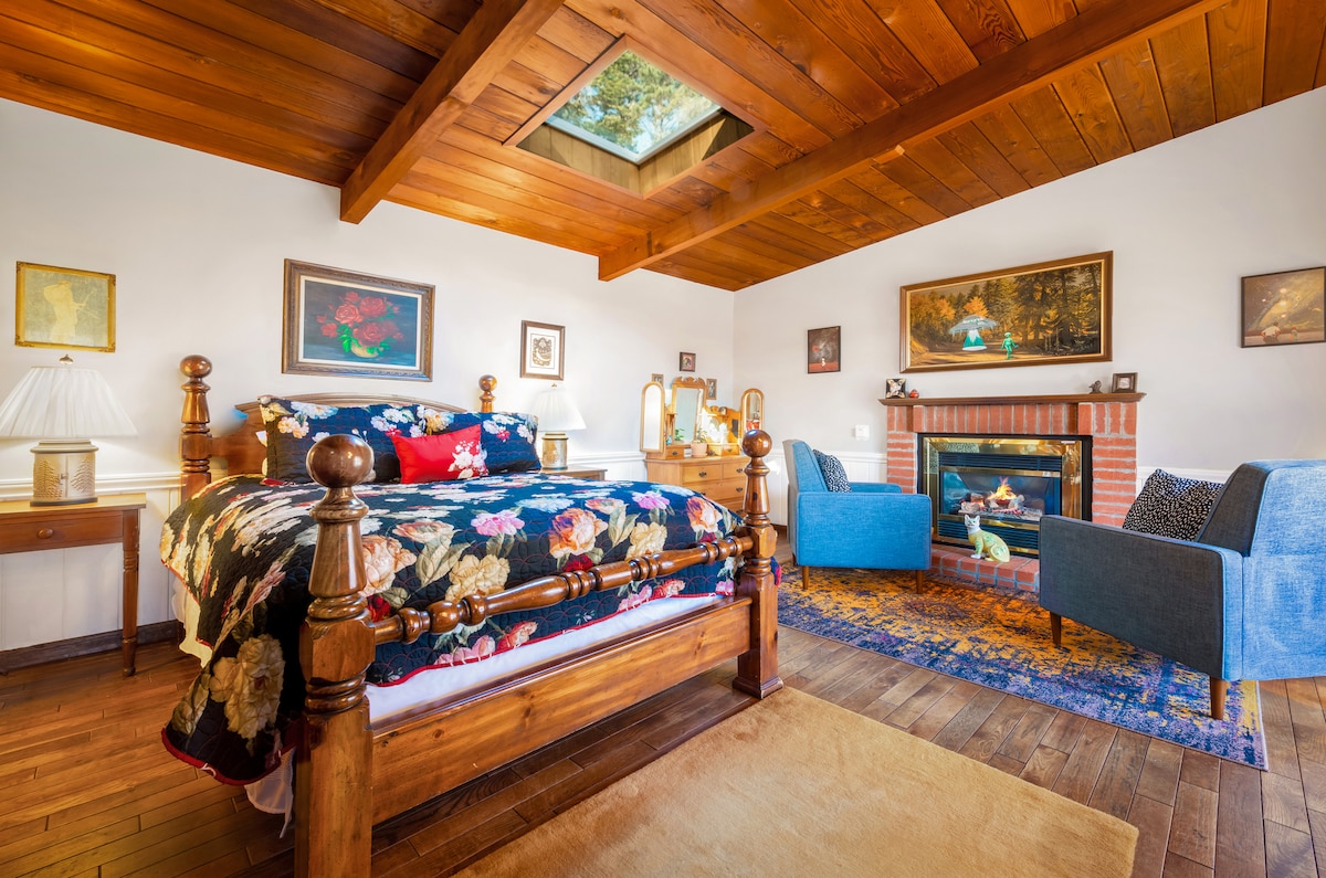 A cozy bedroom features a queen bed adorned with a floral quilt. Two blue chairs are positioned near a charming fireplace, and various artworks hang on the walls. A skylight above allows natural light to illuminate the wooden ceiling and floor.