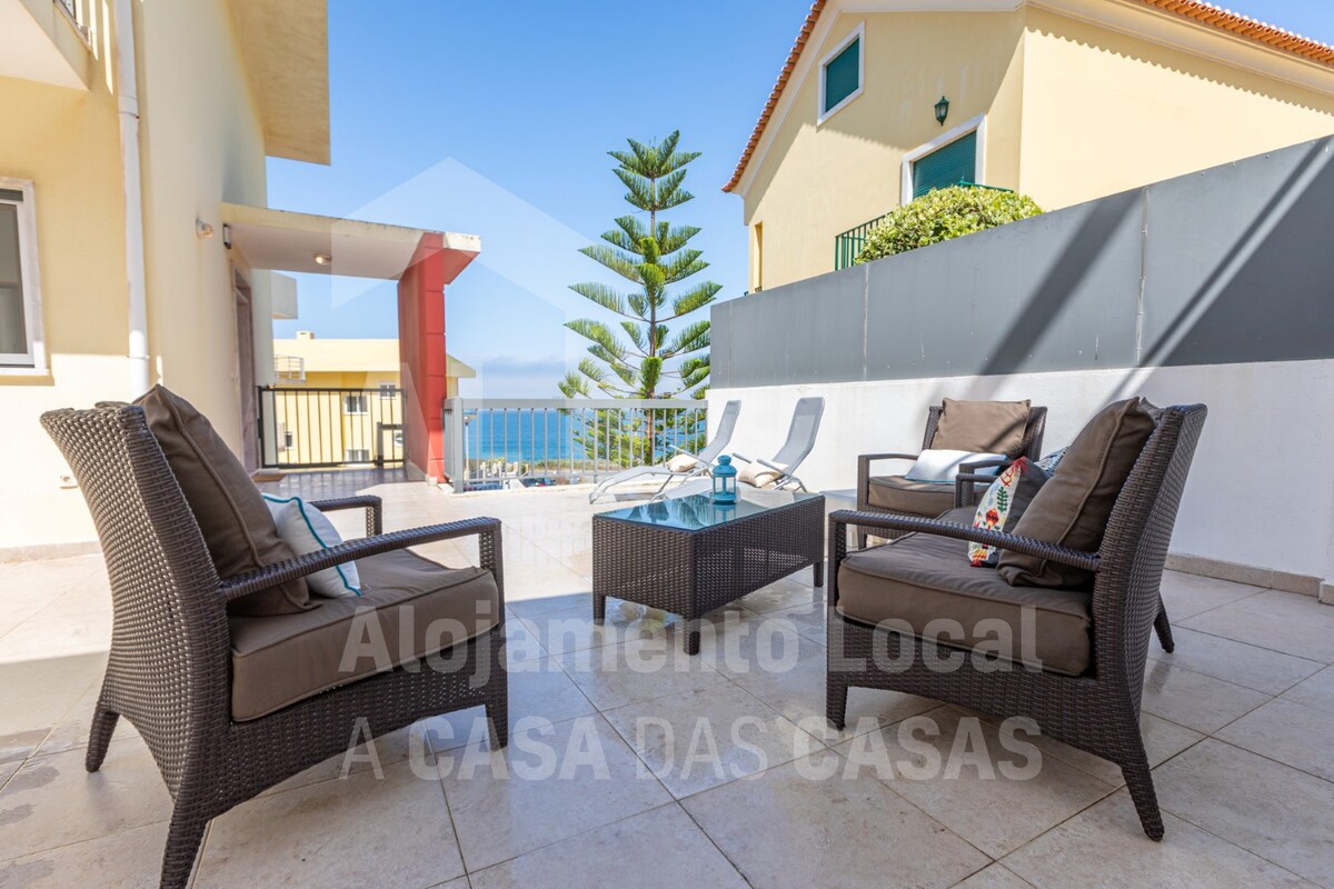 An outdoor terrace area is presented with comfortable seating arrangements. Two loungers are positioned for sunbathing beside a small table centrally located among four cushioned chairs. A view of the sea is framed by the surrounding walls, creating an intimate atmosphere.