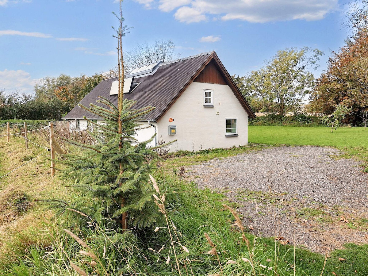 The exterior of a renovated cottage is depicted, featuring a pitched roof and several windows. A grassy area surrounds the home, with a small tree near the corner. A gravel parking space is visible, set against a backdrop of trees and open sky.