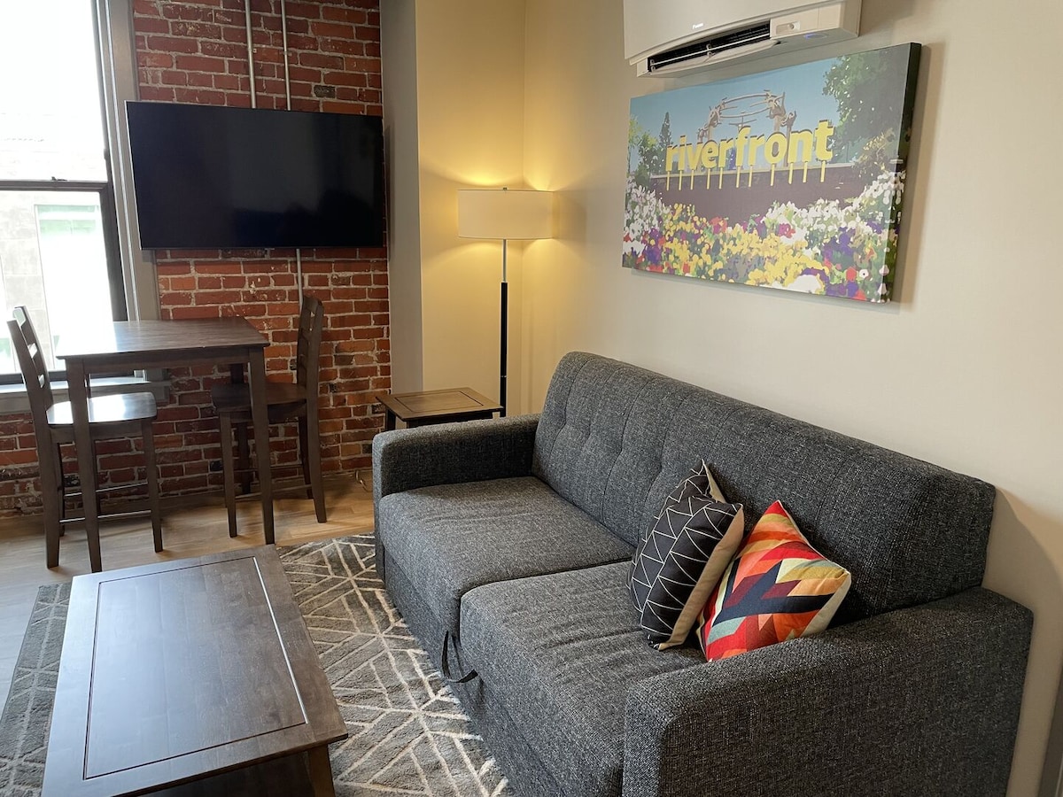 A cozy living area features a gray sectional couch adorned with decorative pillows. A wooden coffee table sits in front, and a dining table with chairs is visible in the background. Exposed brick walls add warmth, complemented by a colorful wall art piece and a flat-screen TV.