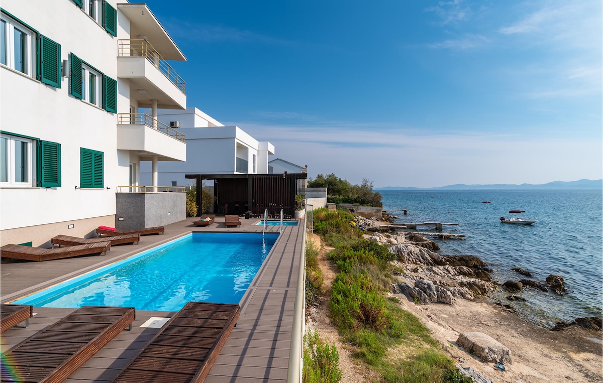 An outdoor private pool is bordered by sun loungers, perfect for relaxation. The clear sea is visible just beyond the pool area, with a rocky shoreline and boats gently anchored in the distance. A bright sky complements the serene coastal setting.