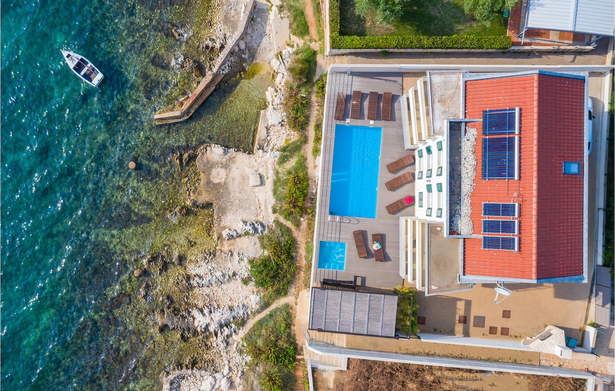 An aerial view captures a vacation home positioned near the clear sea. A private pool, surrounded by sun loungers, is visible alongside stone pathways leading to the water's edge. The red-tiled roof contrasts with the surrounding greenery and rocky shoreline.