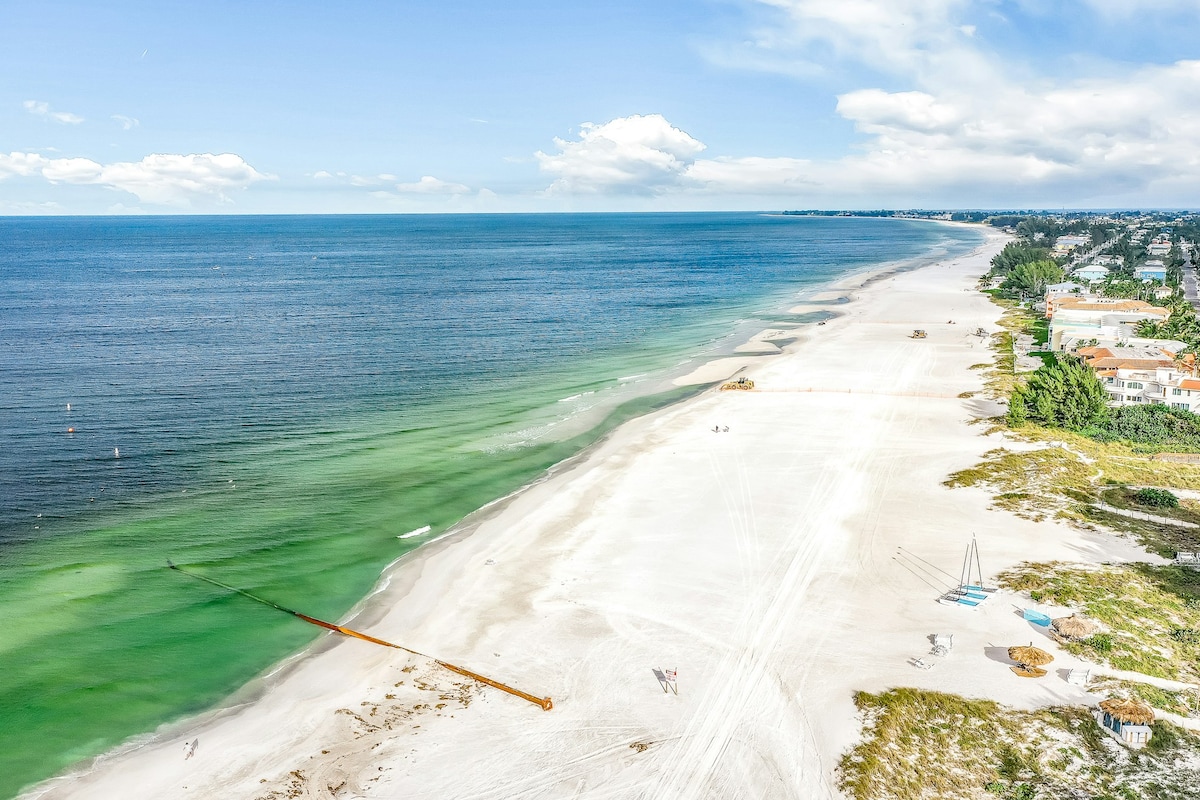An expansive beach stretches out under a clear blue sky, bordered by gentle waves and soft white sand. The shoreline is punctuated by distant beachgoers and lounge chairs, with lush greenery and residences visible along the edge, creating a serene coastal landscape.