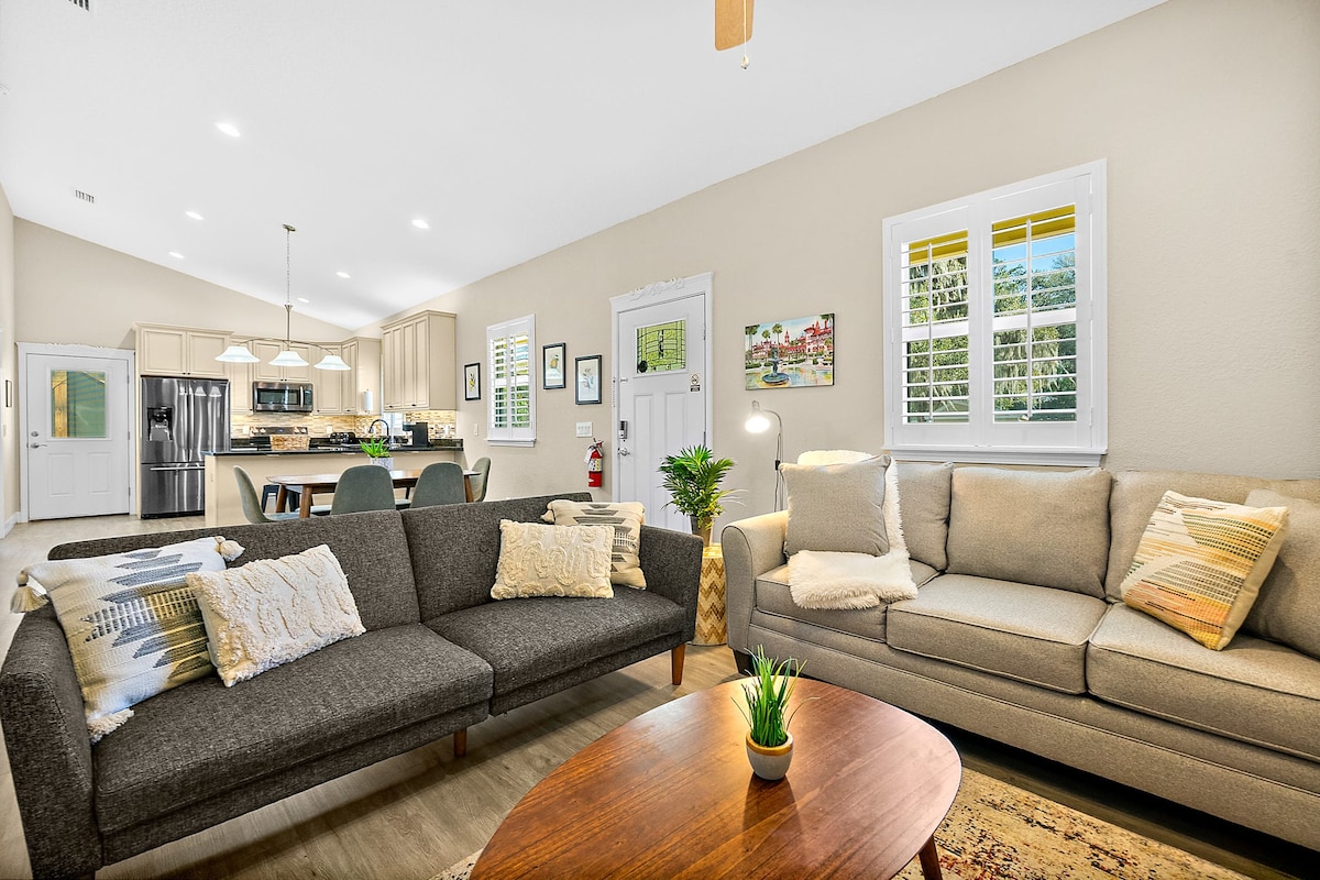 A spacious living area is showcased, featuring two comfortable sofas, a wooden coffee table, and a decorative plant. Bright light filters through the windows, illuminating the neutral-colored walls and creating a warm ambiance. The open layout leads to a well-equipped kitchen in the background.
