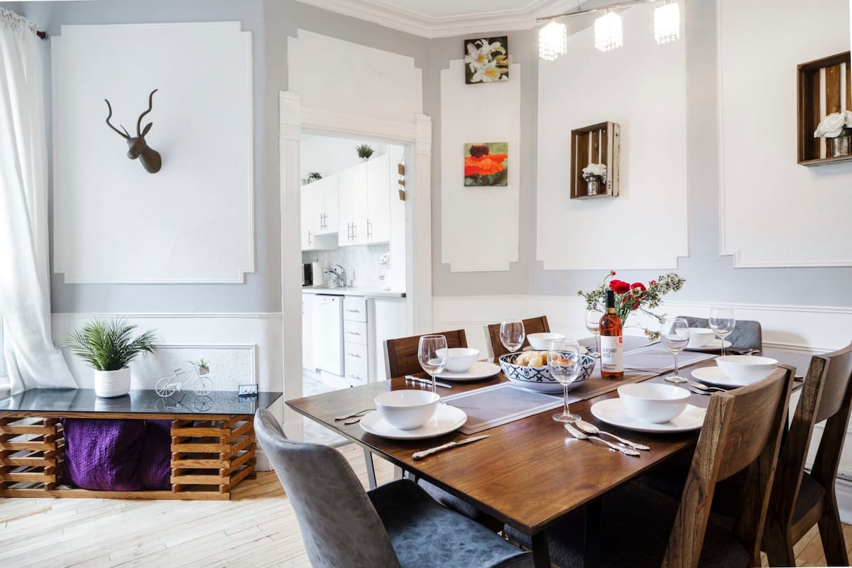A dining area features a wooden table set for six, with white bowls and wine glasses arranged neatly. Decorative wall art adds interest to gray walls, and a sleek side table is adorned with a potted plant. A doorway leads to the kitchen, enhancing the open feel.