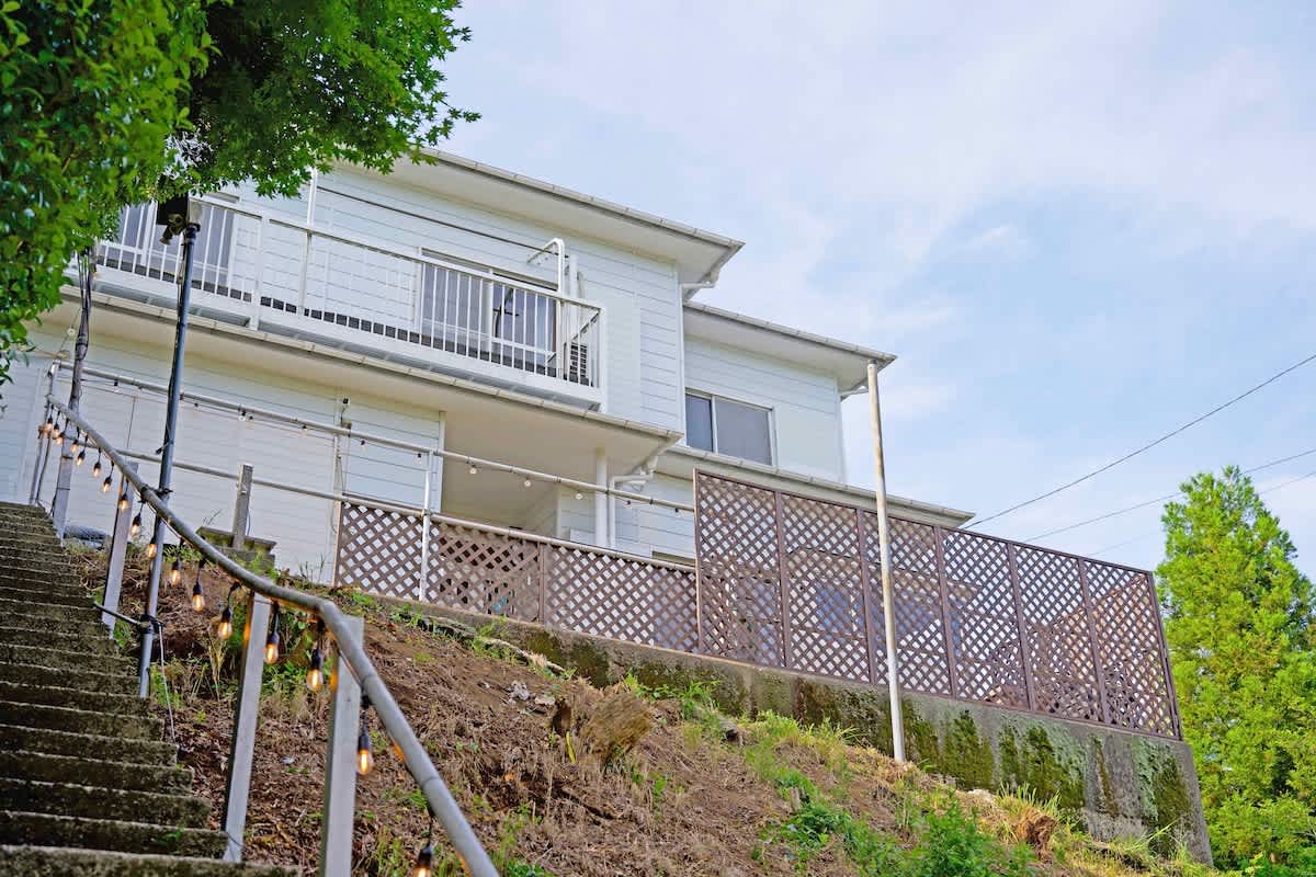 The exterior of a two-story white house is visible from an upward angle, showcasing a spacious balcony with railings. A staircase leads to the entrance, framed by greenery on both sides, and a wooden lattice fence enhances the outdoor area.
