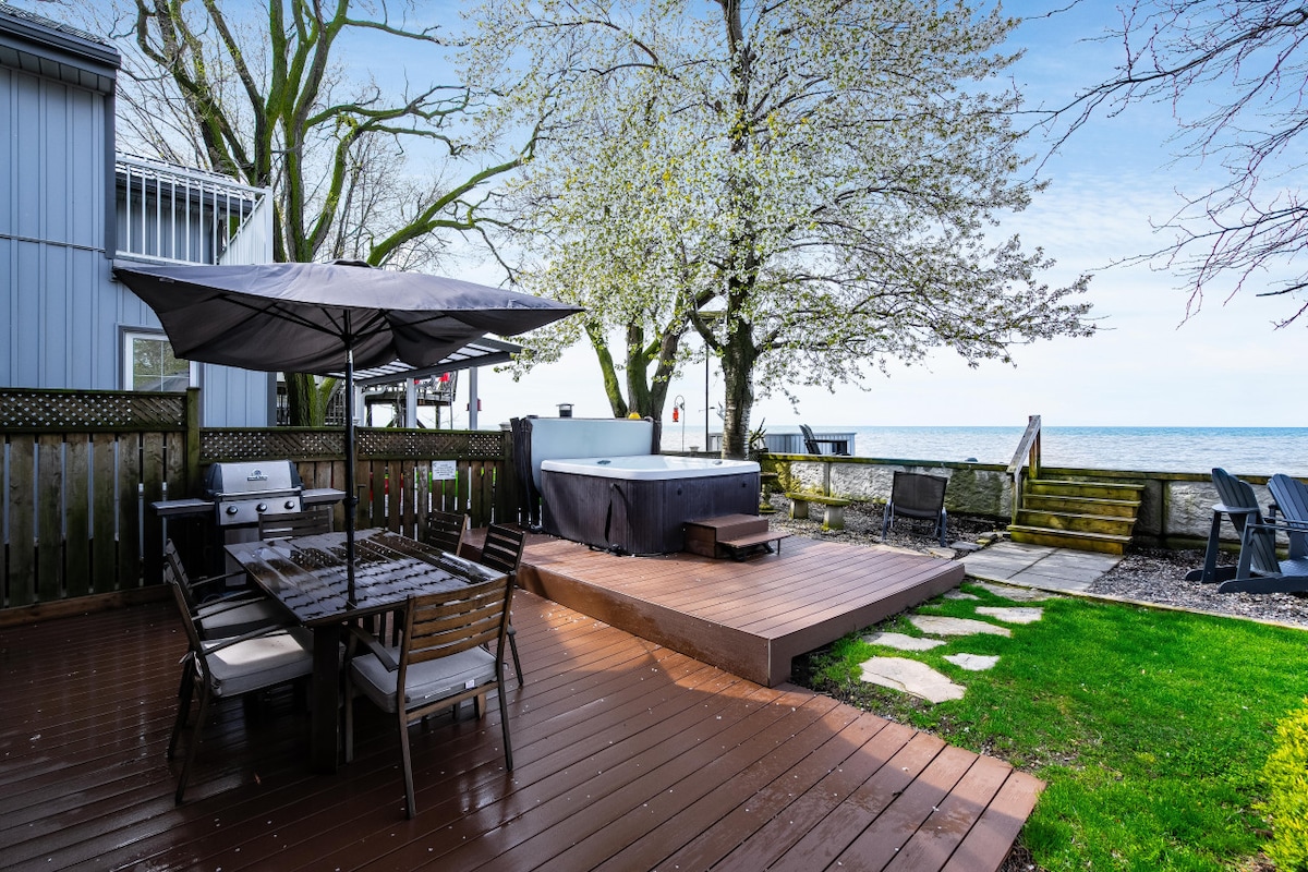 An outdoor patio area features a hot tub surrounded by trees, enhancing the natural setting. A dining table with chairs is placed nearby, accompanied by a barbecue grill. Wooden decking leads to the water, framed by a neat lawn and stepping stones.