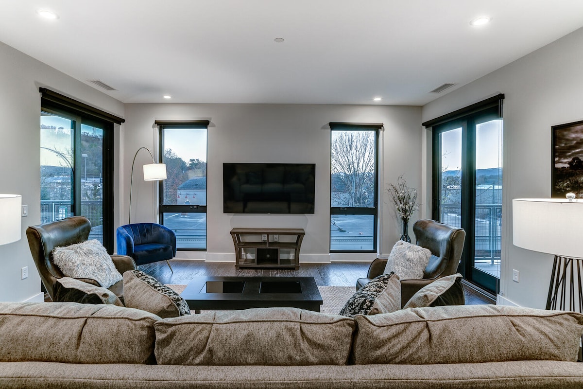 A modern living area is showcased, with large windows allowing natural light to illuminate the space. Comfortable seating, including two armchairs and a spacious sofa, is arranged around a TV stand. Subtle decor elements create a welcoming environment.