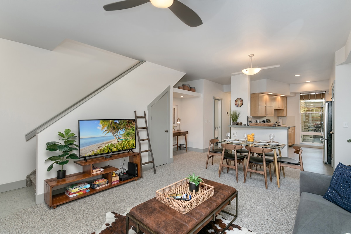 A bright living space is presented with a soft carpet and a comfortable seating area. A large television sits on a media console, while a dining table for six is positioned nearby. Natural light enters through a glass door that leads to the patio area.