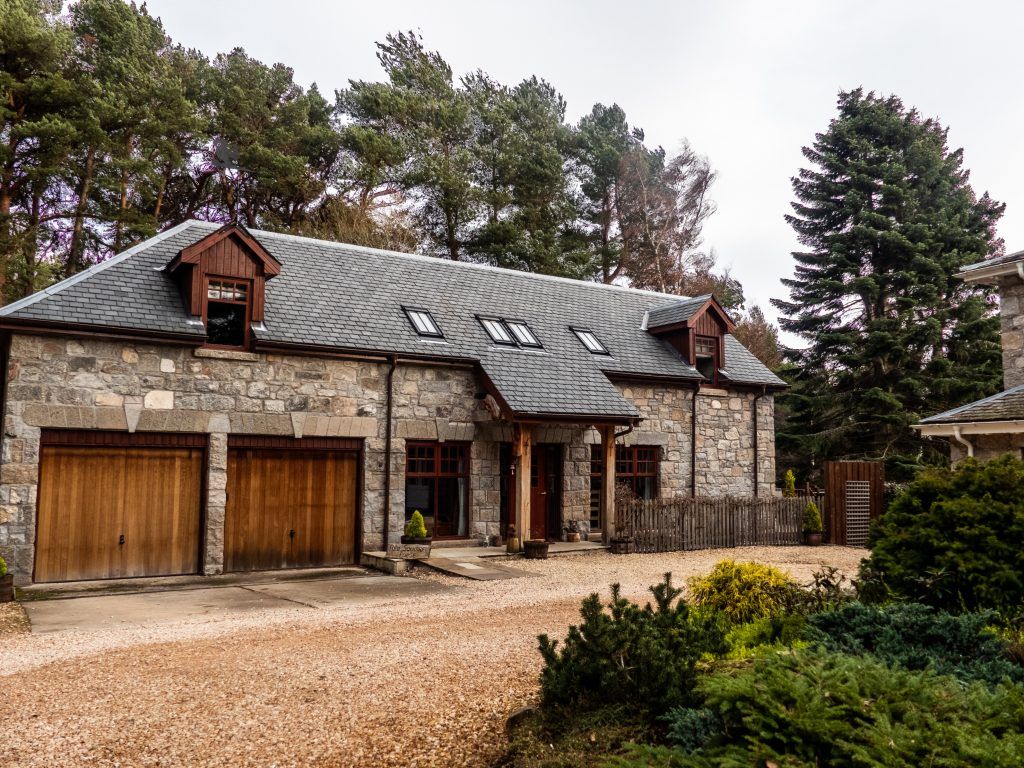 A spacious stone lodge is set amid a tree-lined landscape, featuring a sloping roof and wooden accents. The front entrance is framed by a covered porch, and two garage doors are visible on the ground level. A gravel driveway leads to the entrance.