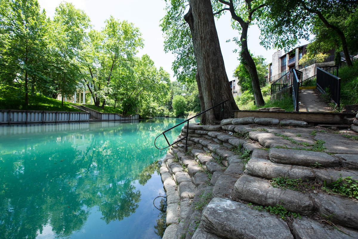 The image displays a clear, tranquil section of the Comal River, bordered by stepped stone pathways leading to the water. Lush greenery surrounds the area, and a tree is positioned nearby, casting a gentle shade. Visible in the distance are condo buildings, enhancing the natural setting.