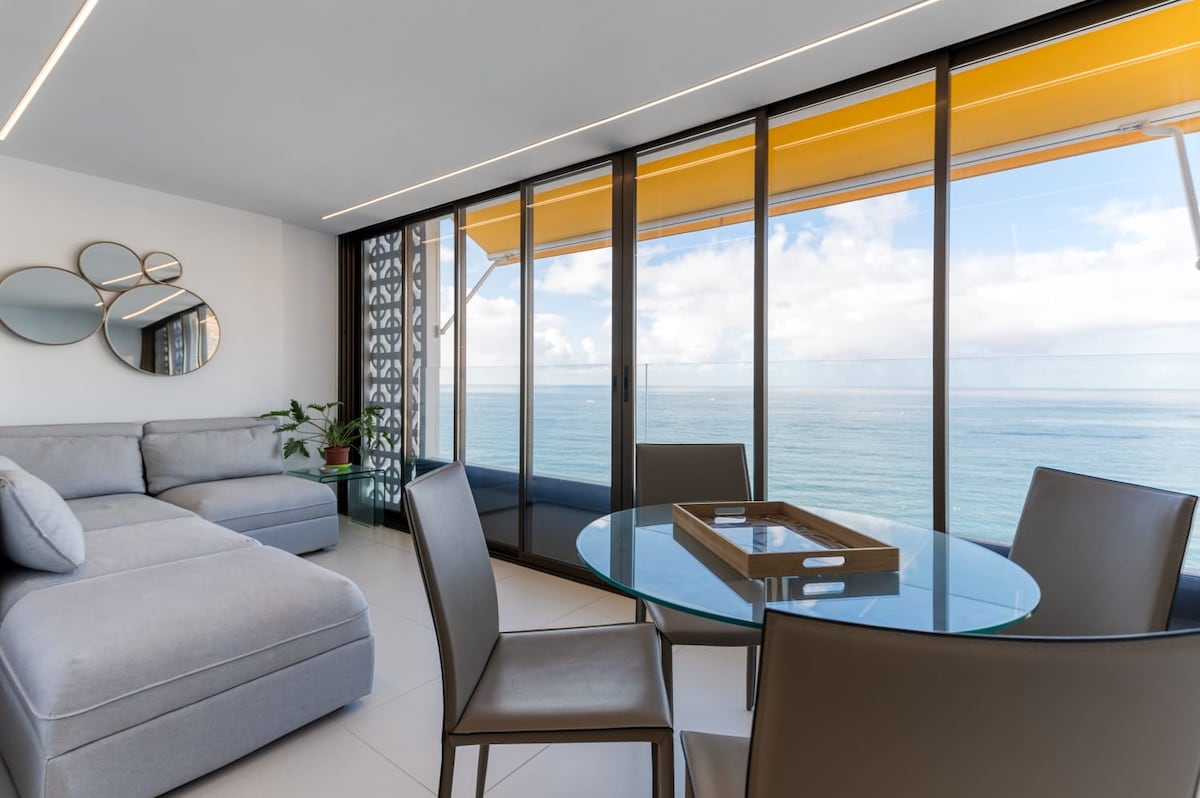 A modern living space is highlighted by large glass doors that reveal sweeping views of the sea. A cozy sectional sofa offers a relaxing area, while a circular glass dining table surrounded by sleek chairs provides an inviting spot for meals. Natural light fills the room.