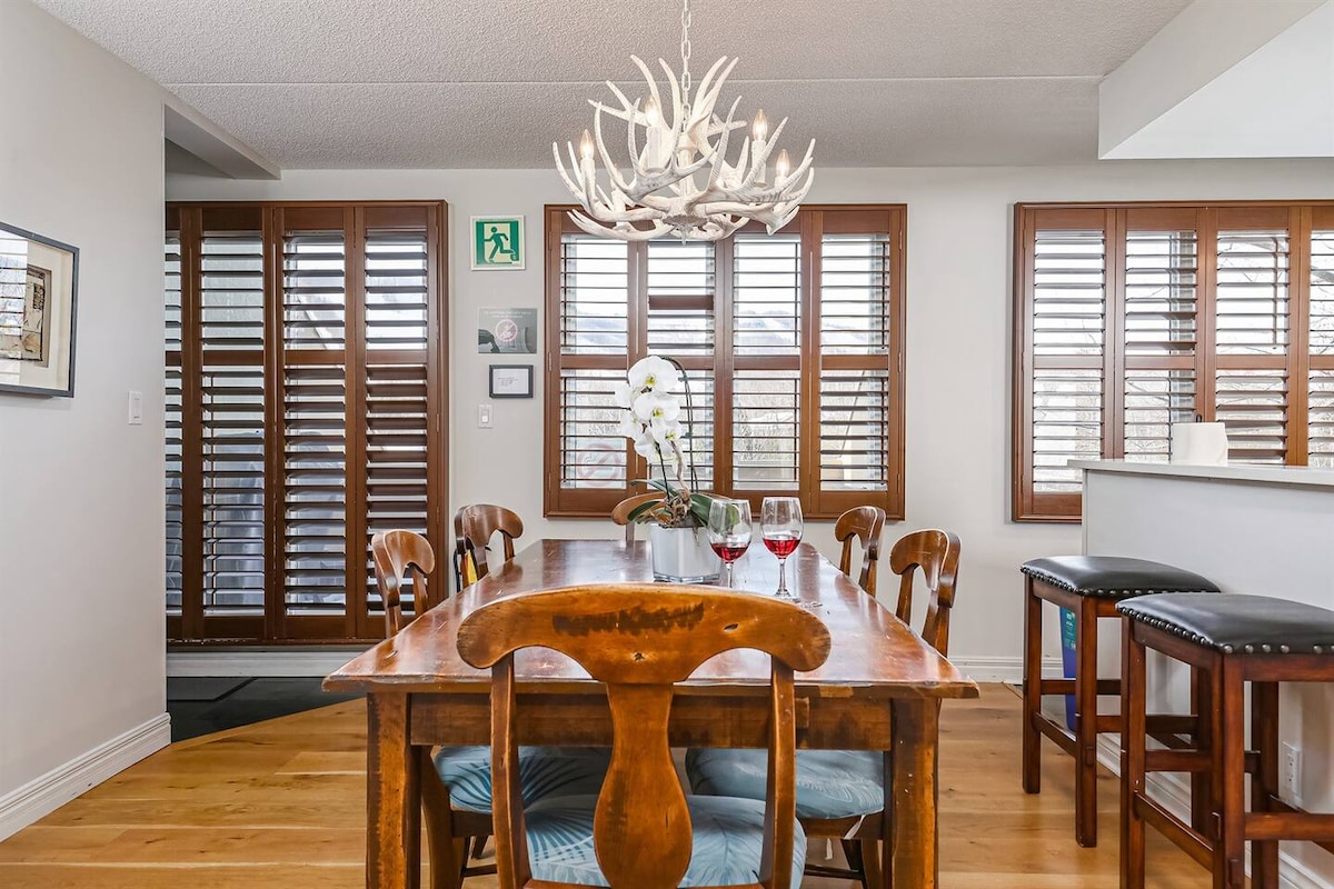A wooden dining table is set for two, complemented by elegant chairs. A chandelier made of antlers hangs above, providing ambient light. Large windows with wooden shutters allow natural light to enter, illuminating the space and highlighting the warm tones of the wooden flooring.