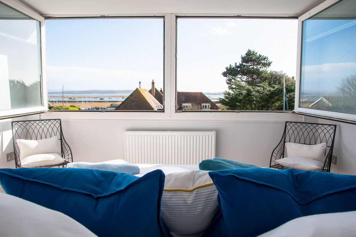 Bright seating area features large picture windows framing expansive views of the Solent and surrounding landscape. Two wire-frame chairs with cushions are positioned to take advantage of the scenery. Soft, navy and cream cushions enhance the inviting space.