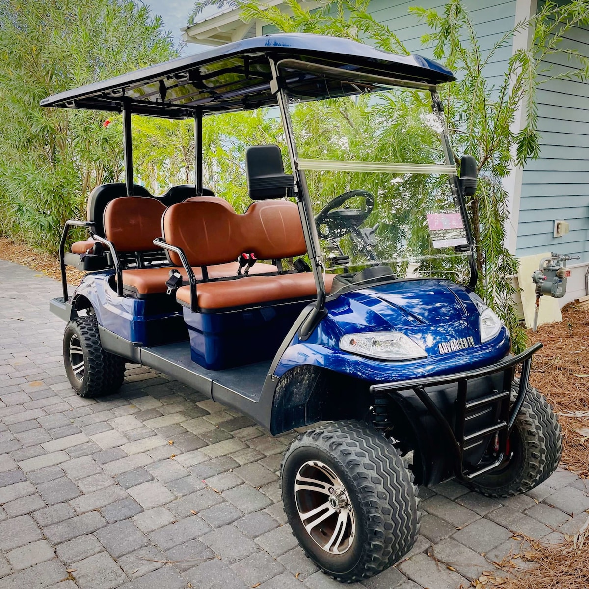 A six-seat golf cart is viewed parked on a patterned stone pathway. The cart features a sleek blue exterior with brown seating and a protective canopy overhead. Lush greenery surrounds the area, contributing to a serene outdoor setting.
