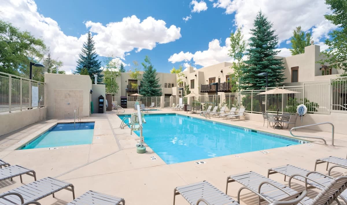 An outdoor pool area features clear blue water surrounded by lounge chairs. Tall trees provide shade, while nearby buildings have balconies overlooking the pool. A separate, smaller pool is also visible to the side, contributing to a relaxed outdoor setting.