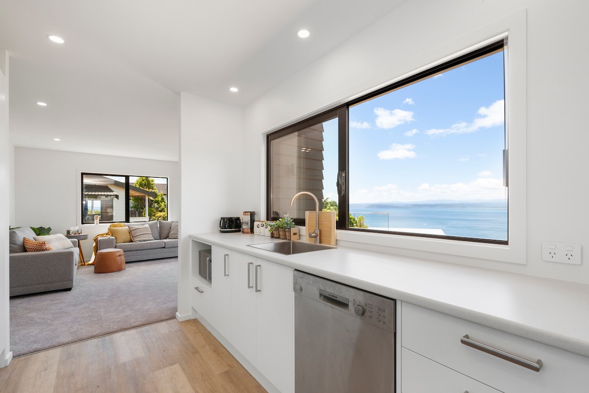 A modern kitchen features sleek cabinetry and a stainless steel dishwasher. Natural light fills the space from a large window that frames a view of the serene lake and sky. Adjacent to the kitchen, a cozy living area can be seen, enhancing the open layout.
