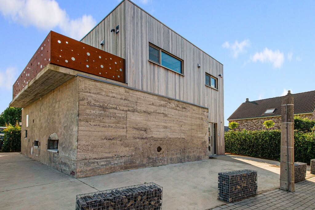 A modern multi-level structure features a blend of materials, including smooth concrete and wooden panels. The upper section showcases a balcony with circular cutouts. The entrance is framed by a minimalist design, while cobblestone seating areas are visible on the ground level.