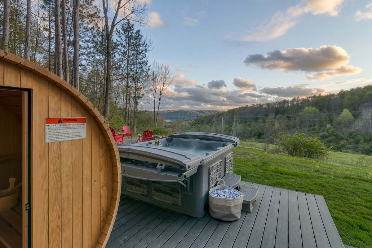 An outdoor area features a barrel sauna adjacent to a hot tub on a wooden deck. Scenic views of rolling hills and trees create a peaceful backdrop, with scattered outdoor seating inviting relaxation.