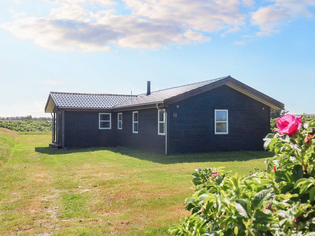 The holiday home is set in a serene environment, featuring a simple yet charming wooden exterior. A grassy area surrounds the house, with vibrant flowers in the foreground. The roof is dark, and a chimney is visible, adding to the property's rustic appeal.