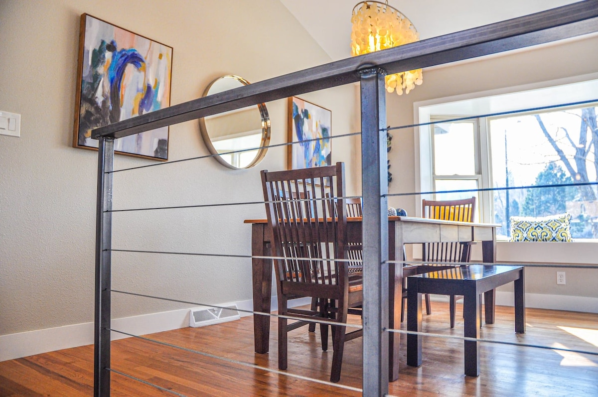 A dining area is highlighted by a wooden table surrounded by four chairs. Artwork decorates the walls, and a round mirror hangs above the table. Natural light enters through a large window, illuminating the space with a warm glow.