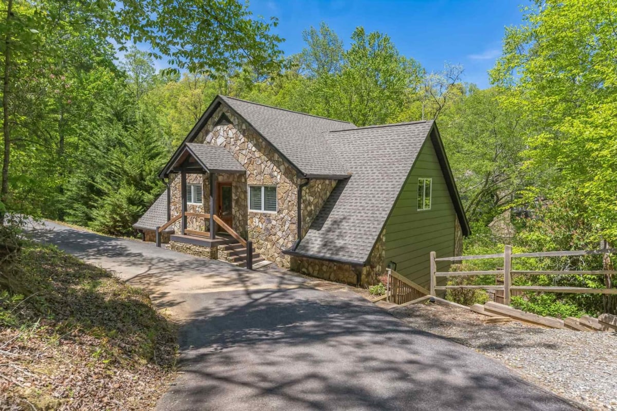 A charming cottage is nestled among lush greenery, showcasing a combination of stone and wood siding. The structure features a steeply pitched roof and large windows that invite natural light. A welcoming path leads to the entrance, enhancing the serene woodland setting.