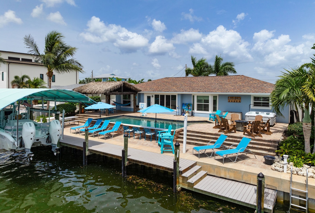 A spacious outdoor area is shown, featuring a large pool surrounded by sun loungers in vibrant colors. A tiki hut with seating is visible, along with a private boat dock. Lush palm trees and blue skies create a relaxed atmosphere.