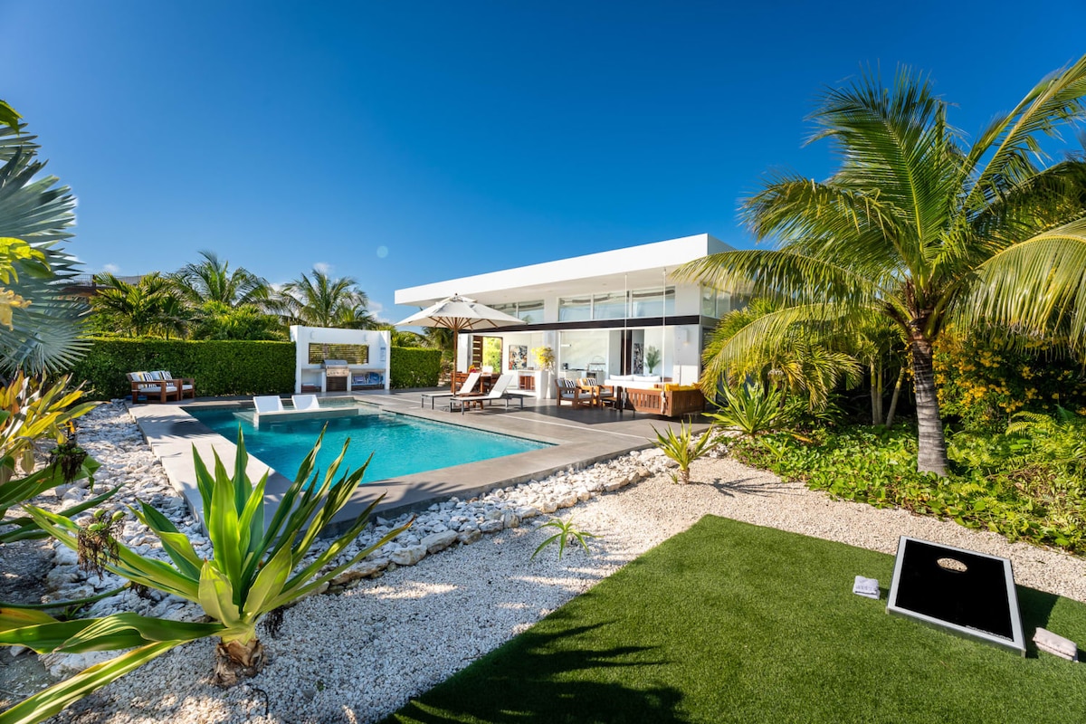A modern villa with a private pool is surrounded by lush tropical landscaping. Sun loungers are positioned near the pool, and a shaded dining area is visible beside it. The outdoor space features a mix of stone and grass, enhancing the relaxed ambiance.