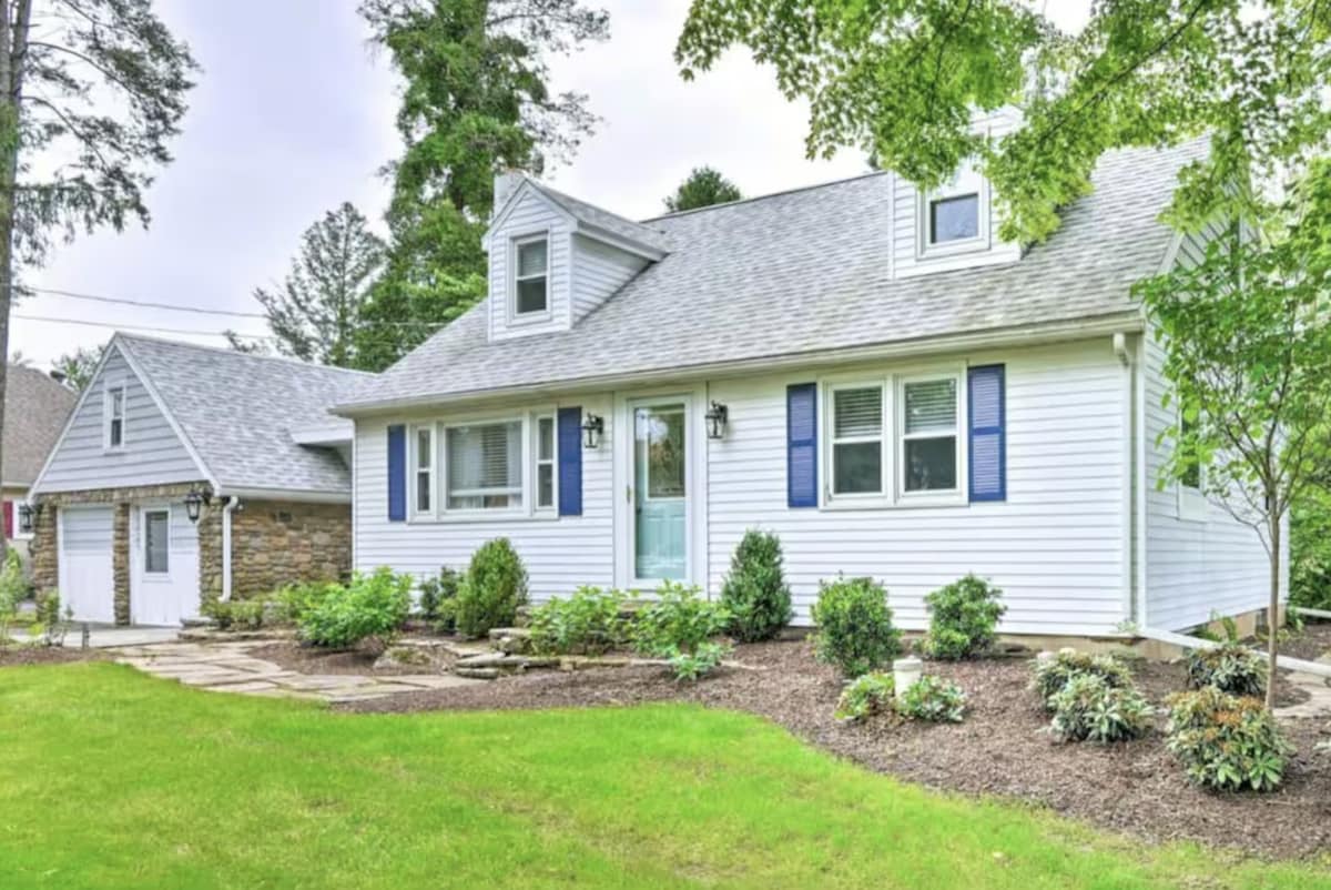 A charming white home features a sturdy gabled roof and blue shutters that complement the light exterior. A stone garage is visible to the left, set against a backdrop of lush green grass and landscaped flower beds, creating an inviting entrance.