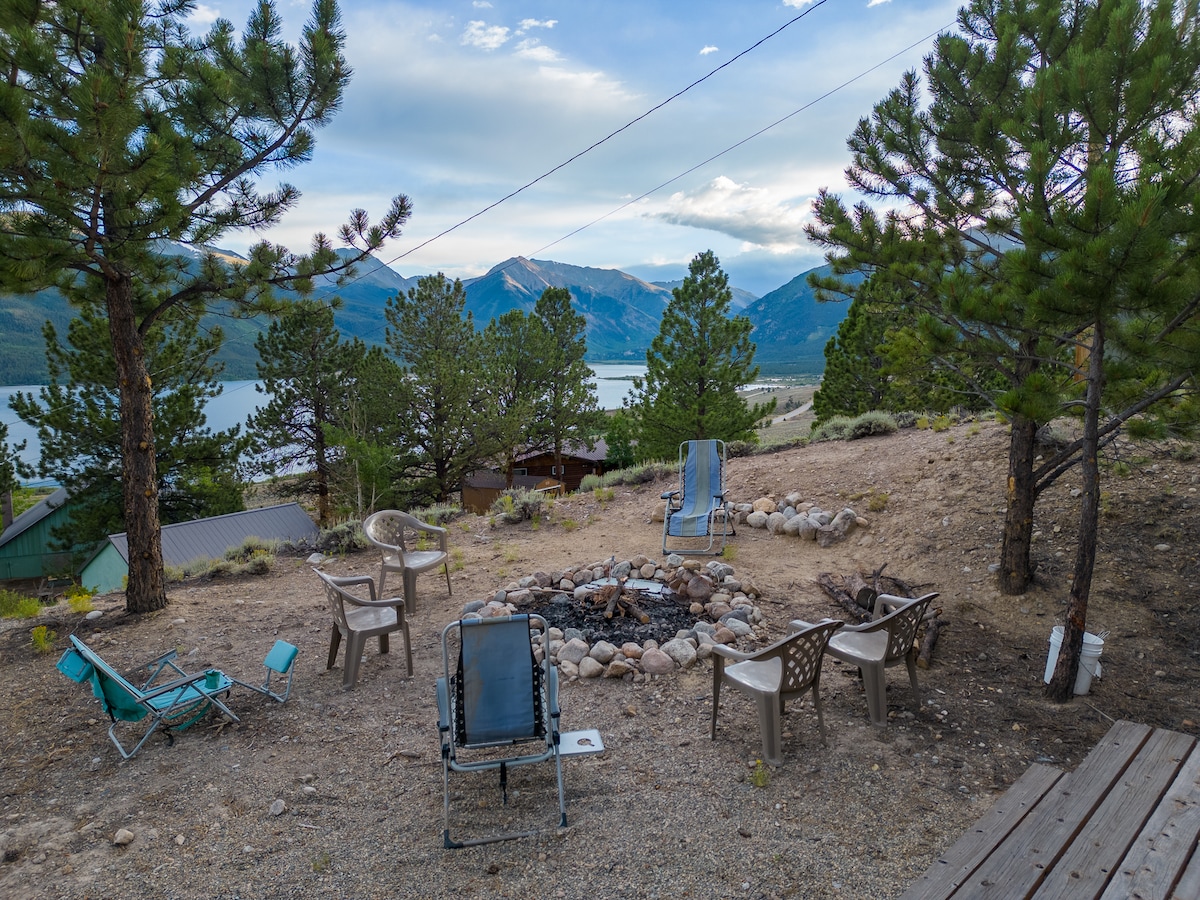 A communal firepit area is surrounded by multiple chairs, inviting relaxation and conversation. The backdrop features serene lake views and majestic mountains, framed by tall pine trees, creating a tranquil outdoor setting.