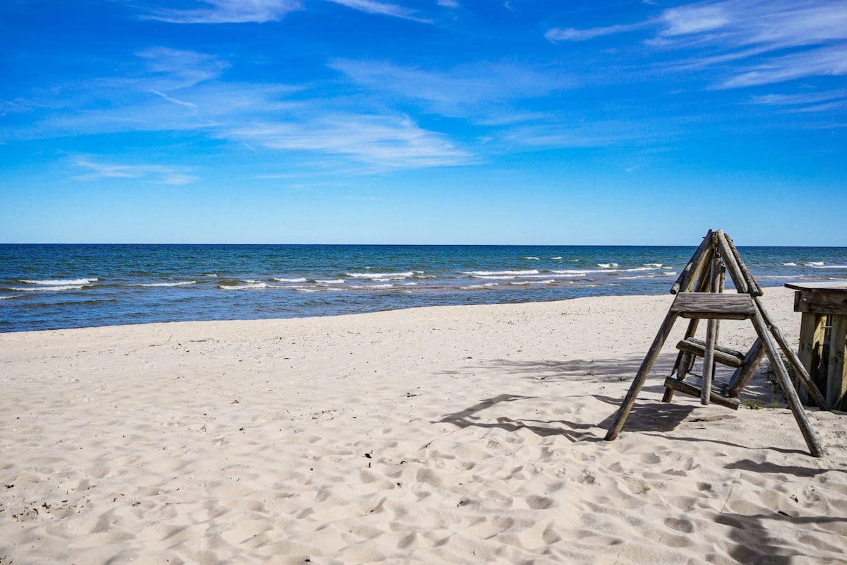 A sandy beach stretches along the coastline, meeting the gentle waves of Lake Huron under a clear blue sky. A wooden swing set is positioned near the shore, offering a playful touch to the serene landscape.